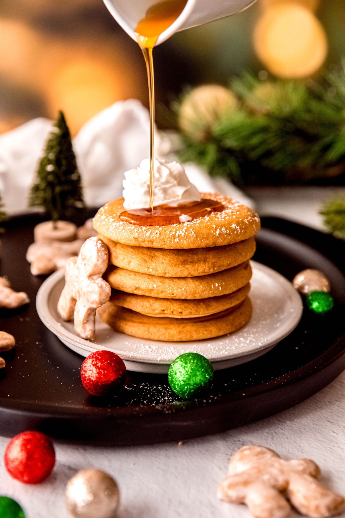 action shot of maple syrup being poured on a stack of gingerbread christmas pancakes with powdered sugar and a squirt of whipped cream with pine branches and fairy lights blurred out in the background.