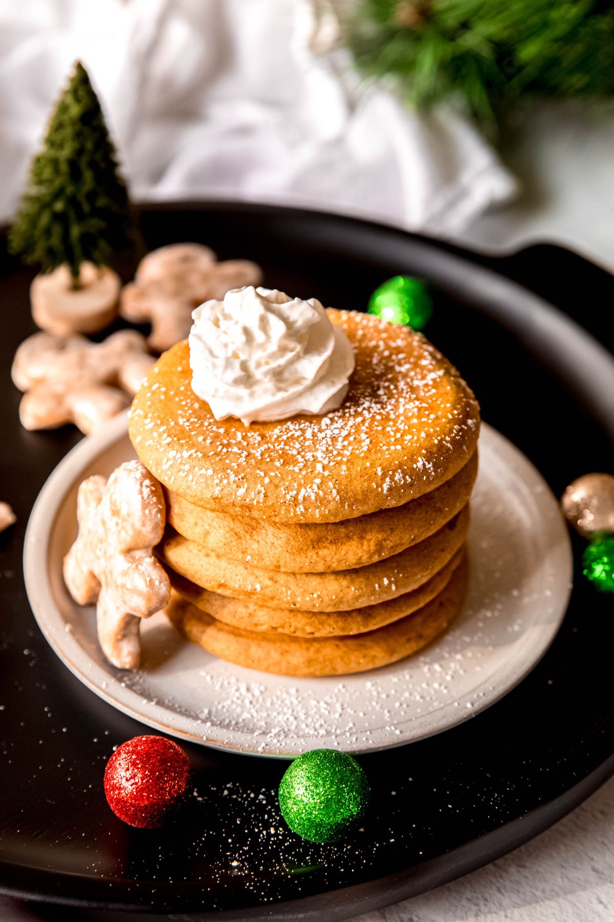 stack of fluffy gingerbread pancakes on a white plate on a black serving platter; the stack is topped with a squirt of whipped cream and dusted with powdered sugar.