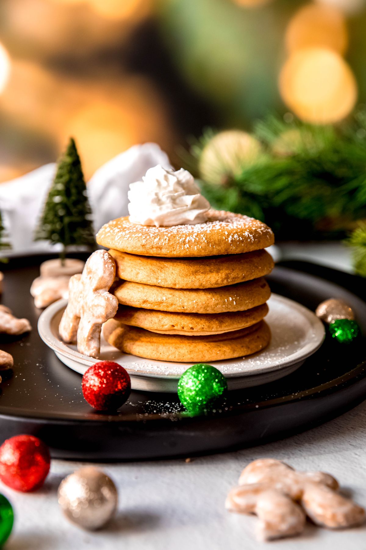 stack of 5 whole wheat gingerbread pancakes on a white plate on a black serving tray with powdered sugar, mini gingerbread men and mini christmas ornaments.
