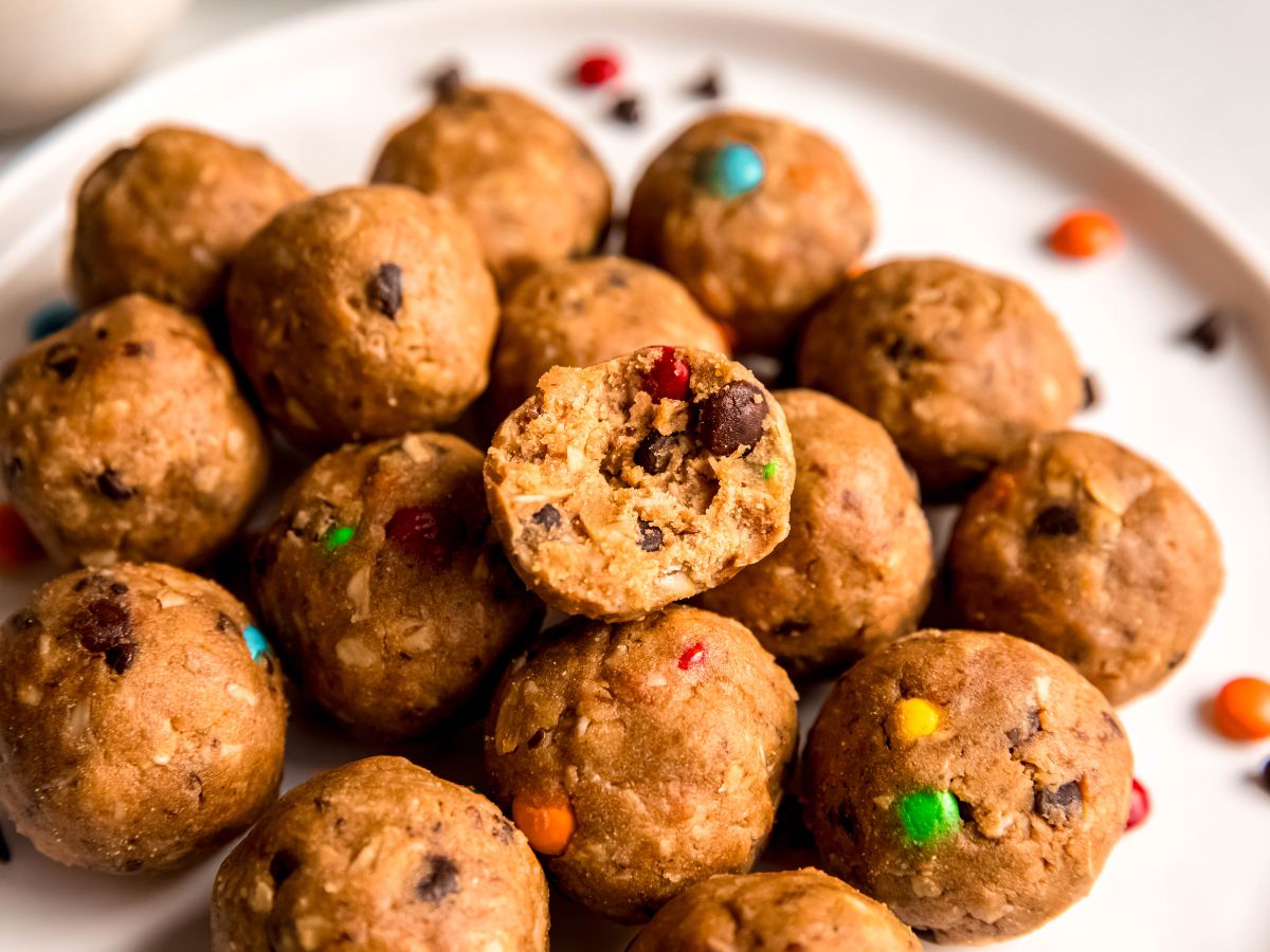 horizontally framed hero shot of a pile of monster cookie energy balls on a plate with the one on top bit in half to show the texture.