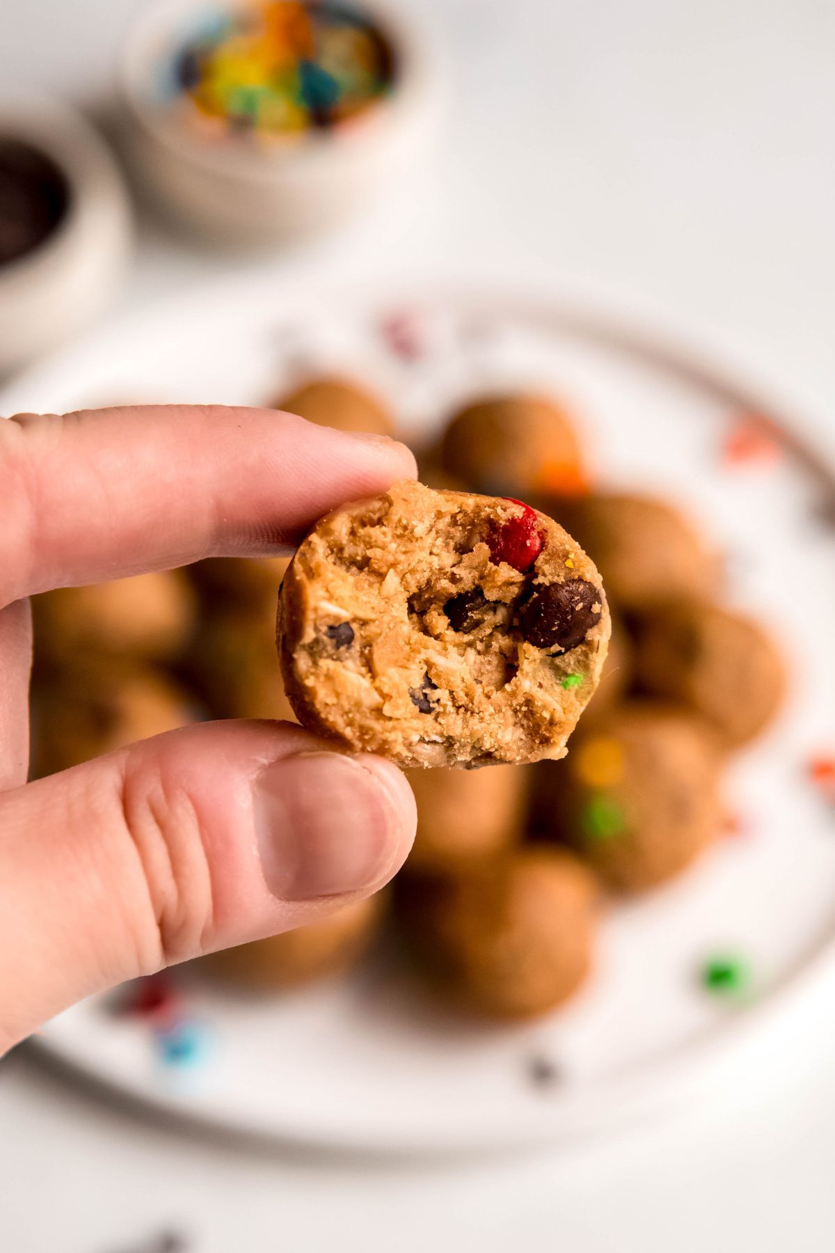 hand holding a monster cookie protein ball that has been bit in half to show the cookie dough-like texture with the rest of the plate blurred out in the background.