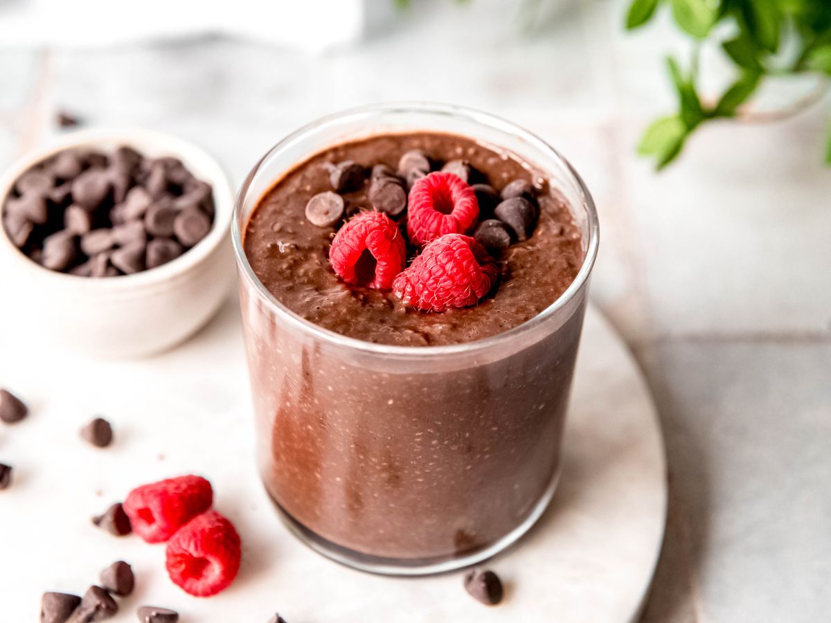horizontally framed hero shot of a garnished glass of chocolate protein chia pudding on a white table with a white bowl of chocolate chips and some raspberries.