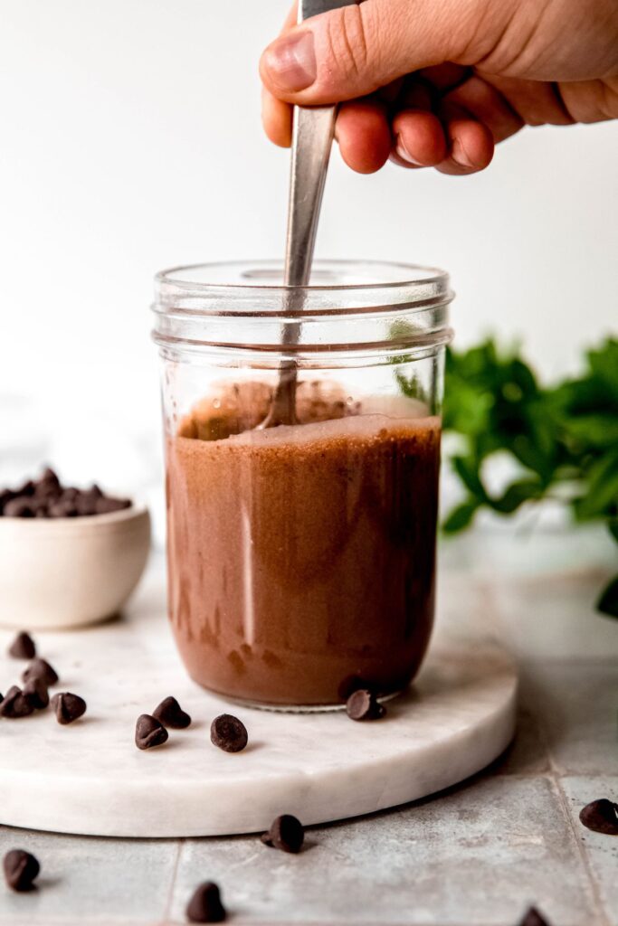 action shot of a hand stirring in chia seeds into the chocolate protein milk mix with a silver spoon.