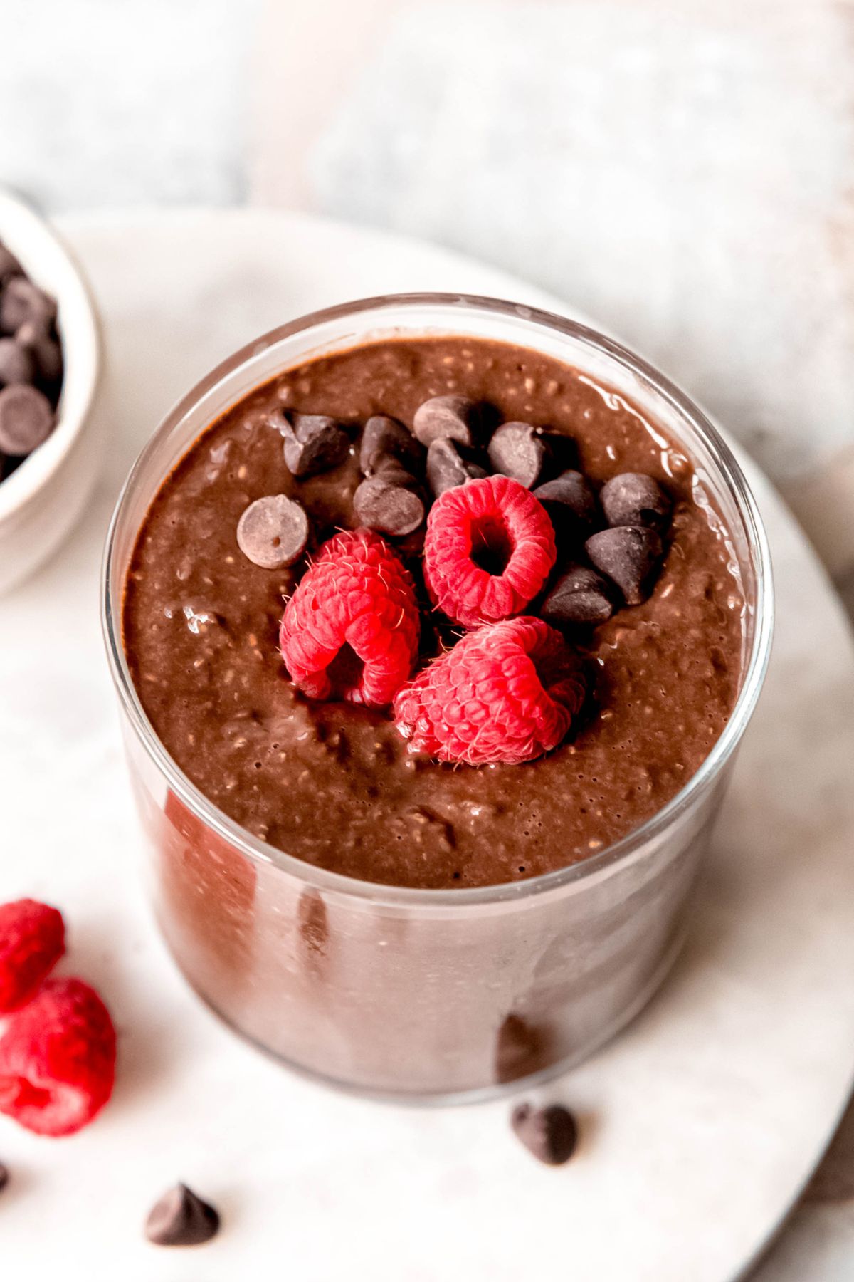 closeup overhead shot of a raspberry and chocolate chip garnished glass of high protein chocolate and chia seed pudding on a white table with more raspberries and chocolate chips scattered around.