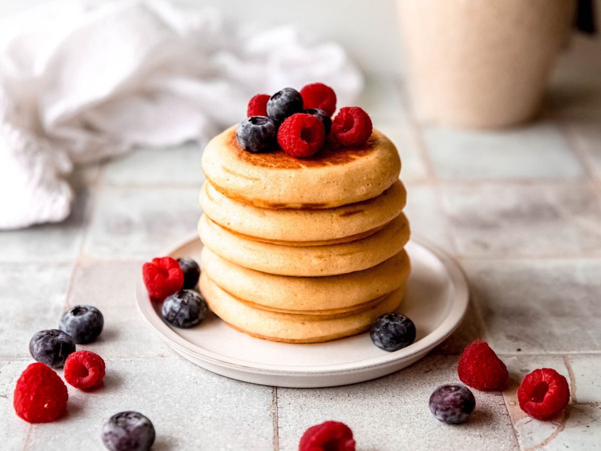 horizontally framed hero shot of a grey tile table with a white plate of fluffy protein pancakes topped with fresh raspberries and blueberries with more berries scattered across the table.