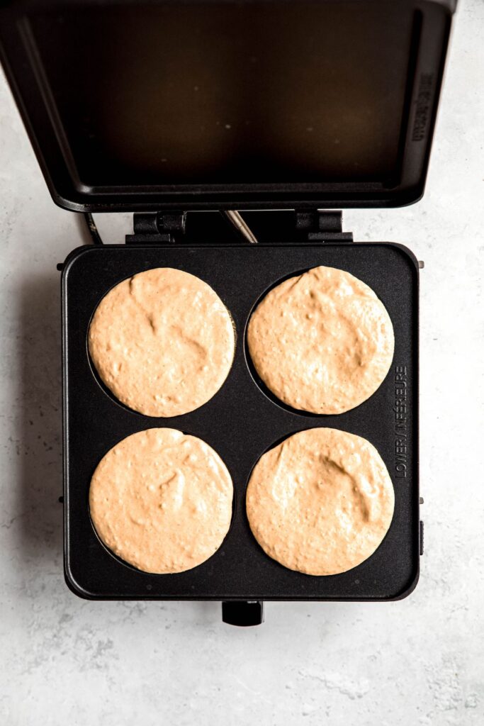 4 raw circles of protein powder pancake batter cooking on a nonstick electric griddle.