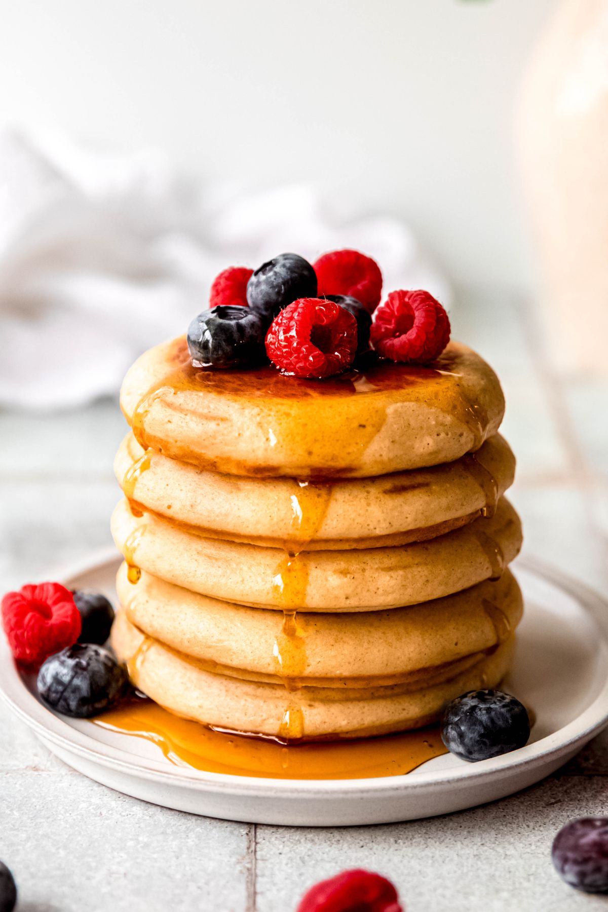 vertically framed hero shot of a stack of protein powder pancakes topped with fresh berries and maple syrup on a white plate on a grey tile table.