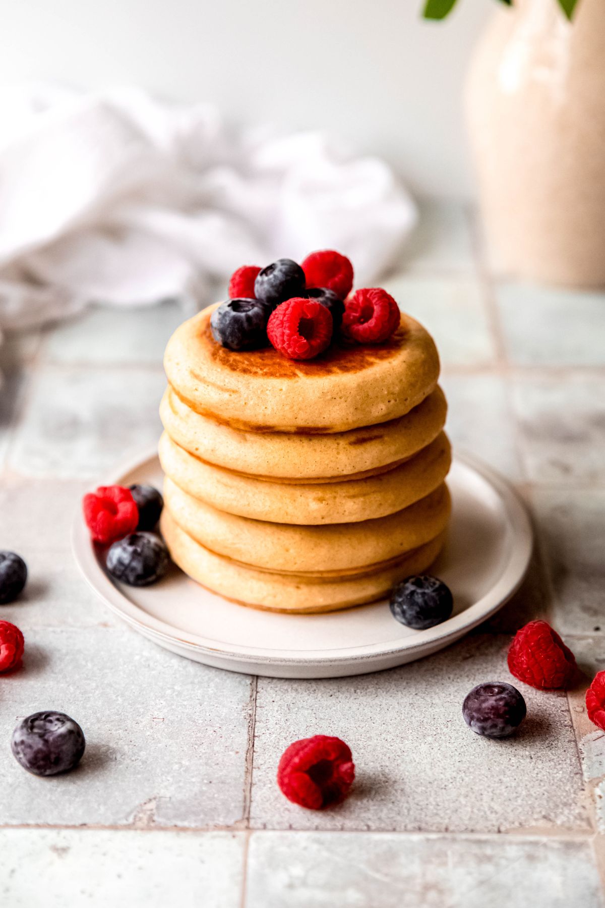 stack of 5 protein powder pancakes on a round white plate topped with fresh blueberries and raspberries; there are some errant berries scattered around on the grey tile table.
