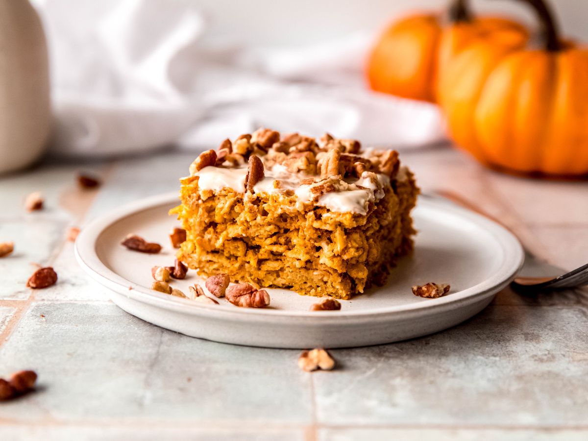 horizontally framed shot of a slice of high protein baked pumpkin pie oatmeal on a white plate on a grey slate tile table with orange pumpkins blurred out in the background and chopped pecans scattered around.