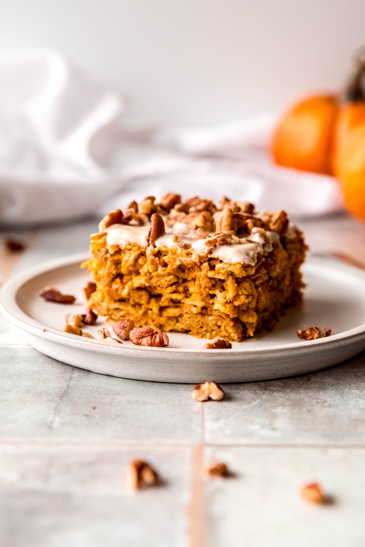 slice of pumpkin baked oatmeal with cream cheese glaze and pecans on a white plate on a grey slate table with orange pumpkins blurred out in the background.