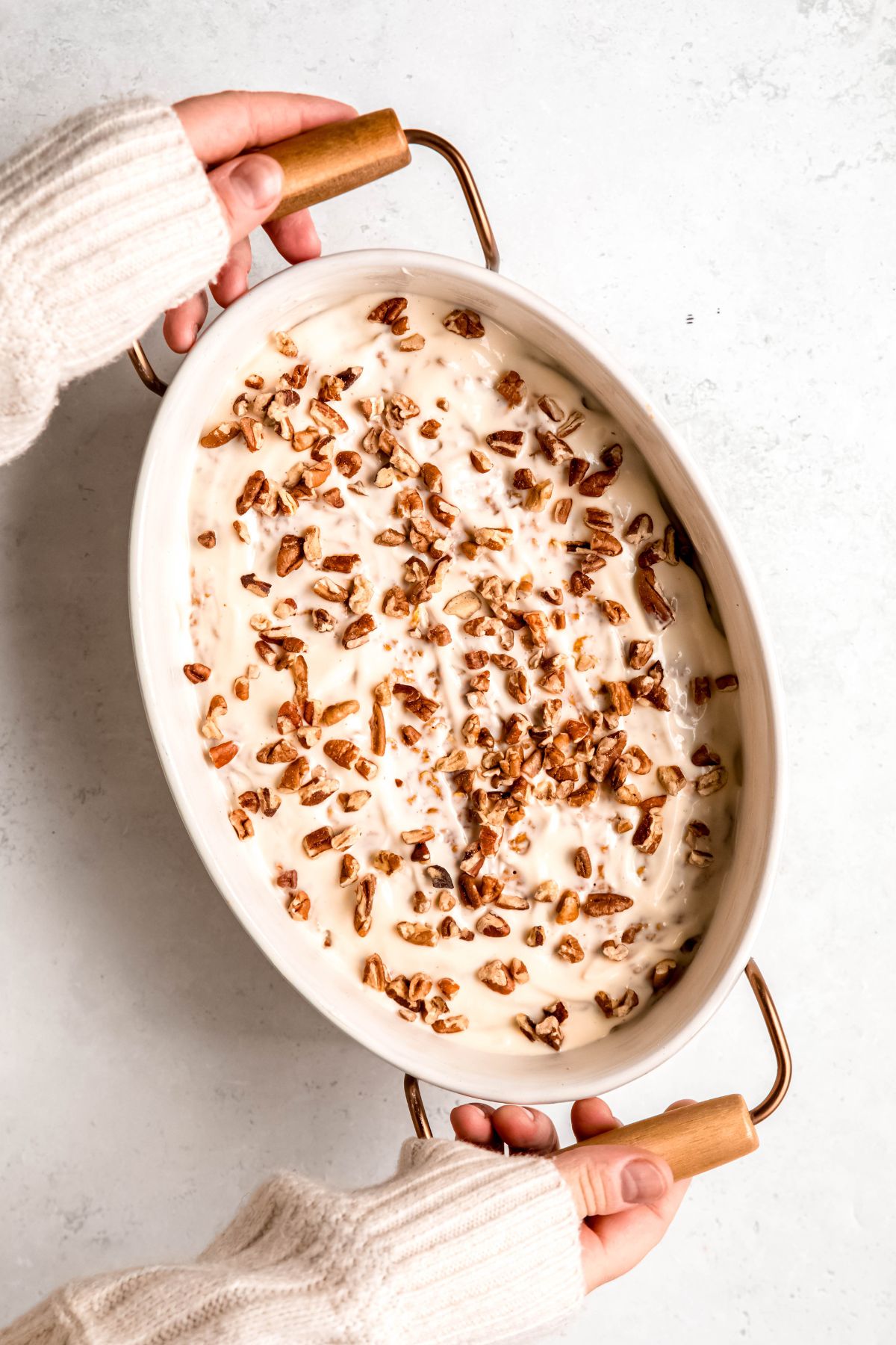 hands clad in a white sweater up to the knuckles using metal and wooden handles to set down a casserole dish of high protein pumpkin baked oatmeal on a white table.
