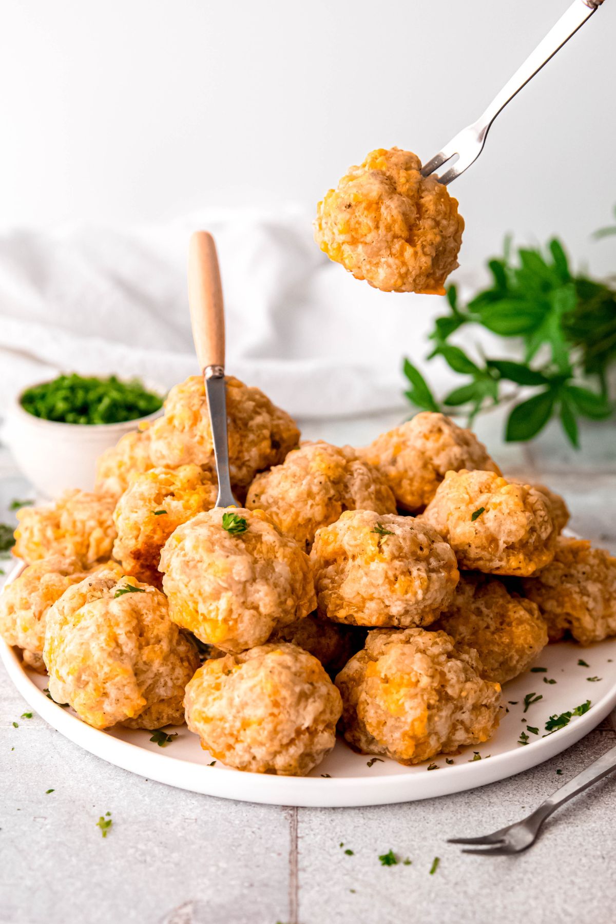shot of a serving platter with a jumble of no bisquick sausage balls with silver and wooden appetizer forks and a pinch bowl of fresh parsley in the background.