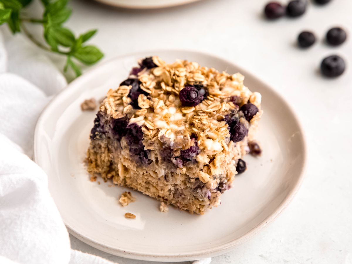 closeup shot of a slice of healthy blueberry baked oatmeal on a white dessert plate on a white table with fresh blueberries.