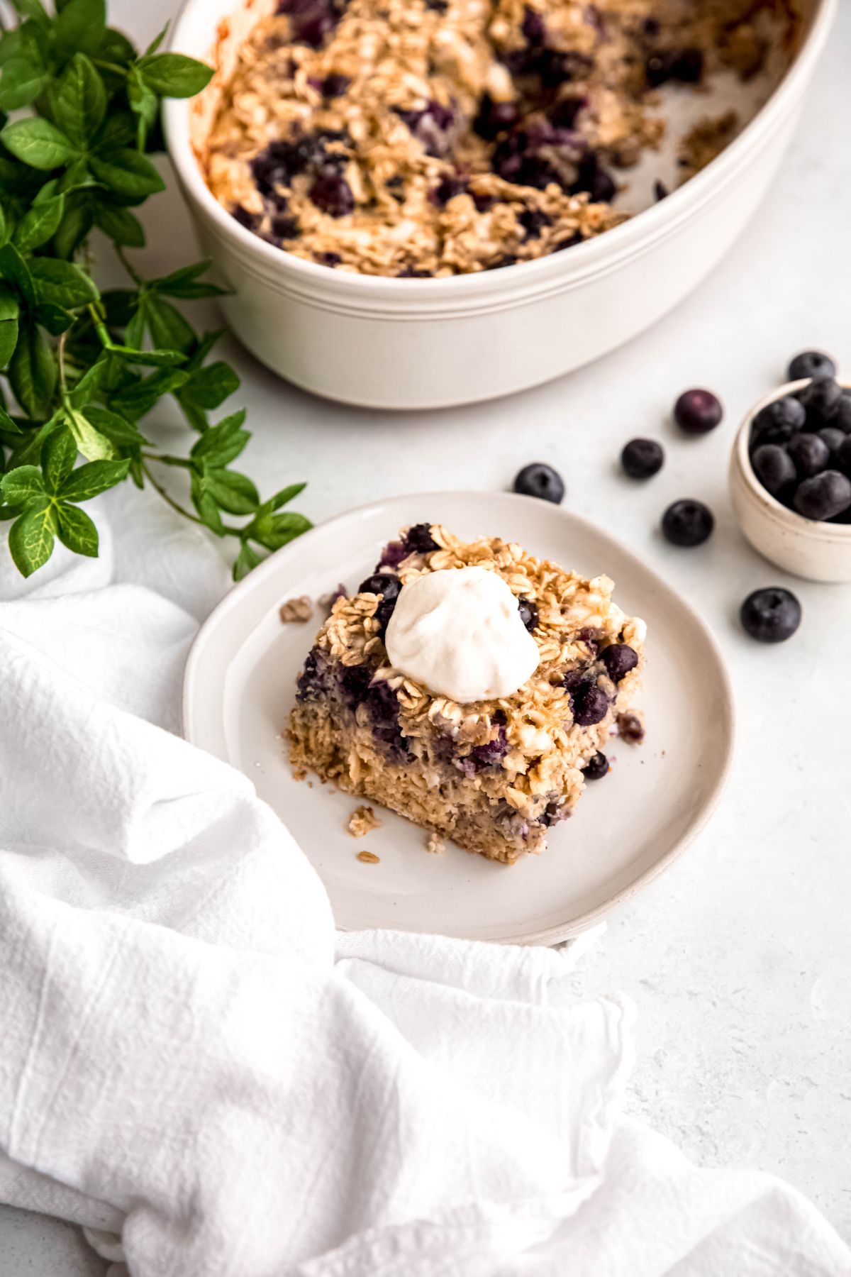 round white dessert plate with a serving of a high protein blueberry oatmeal bake topped with a dollop of yogurt on a white table with fresh blueberries, a white linen cloth, and a green houseplant.