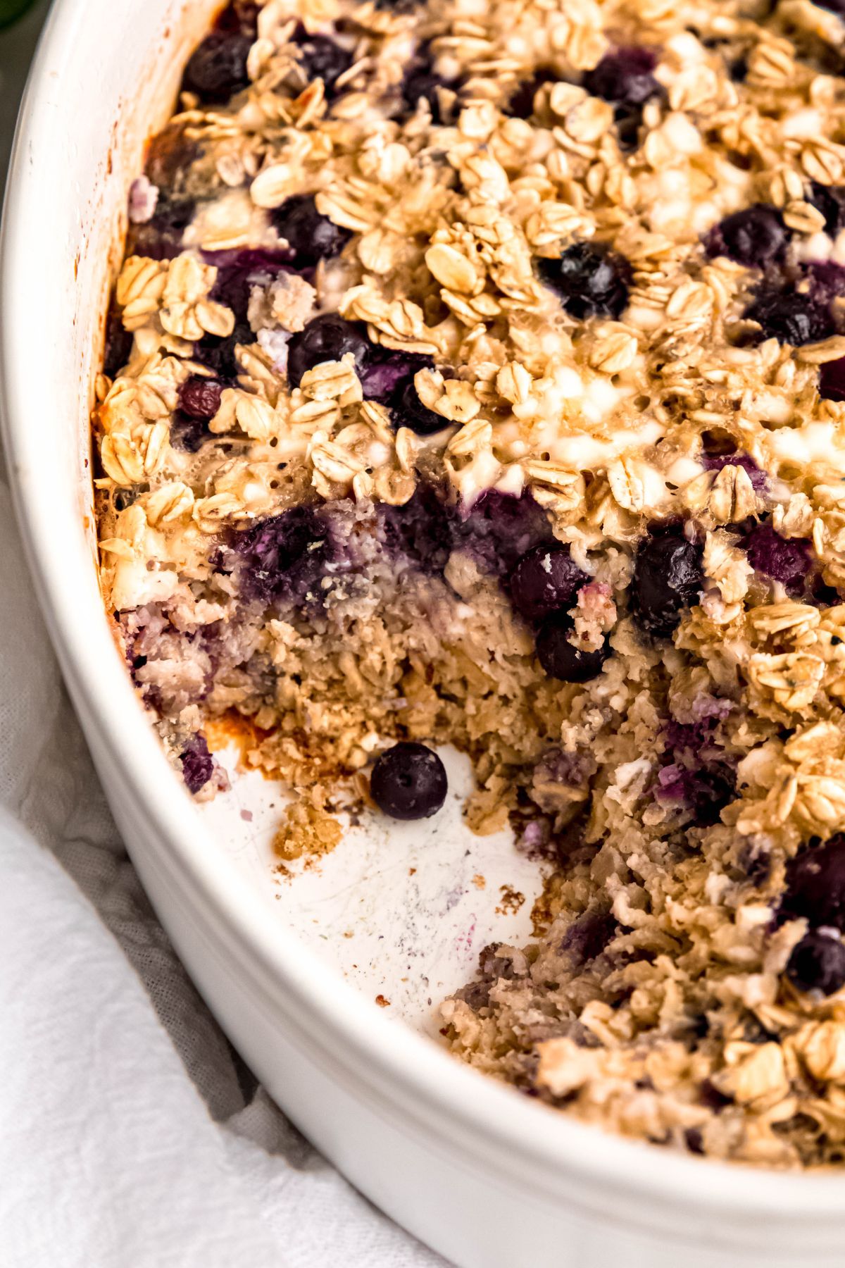 closeup shot on a white casserole dish with blueberry baked oatmeal with a portion removed showing the cake-like texture and juicy blueberries throughout.