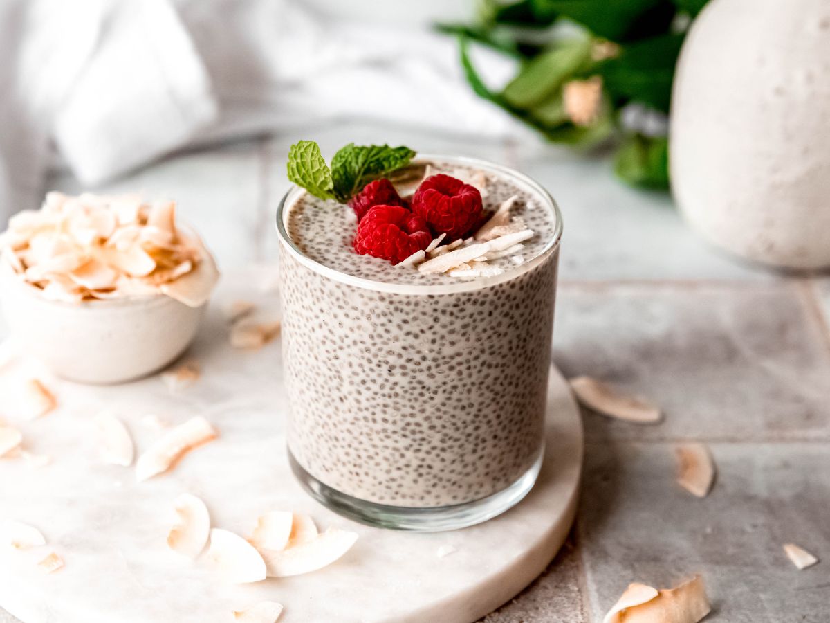 horizontally framed hero shot of a glass of coconut-flavored chia protein pudding on a white marble round on a grey slate table with a pinch bowl of coconut flakes and a green houseplant blurred out in the background.