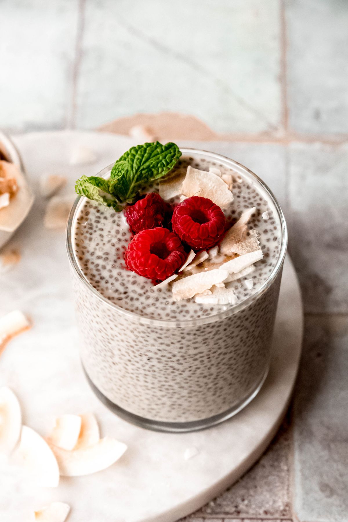 closeup overhead angled shot looking down into a garnished glass of high protein coconut chia seed pudding topped with toasted coconut flakes, raspberries, and a sprig of mint with more coconut flakes scattered on the white table below.