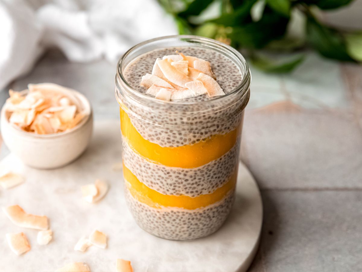 hero image of a coconut mango chia protein pudding parfait on a white marble round on a grey slate table; the parfait is topped with coconut flakes and there is a bowl of extra toasted coconut flakes to the side and a green houseplant in the back.