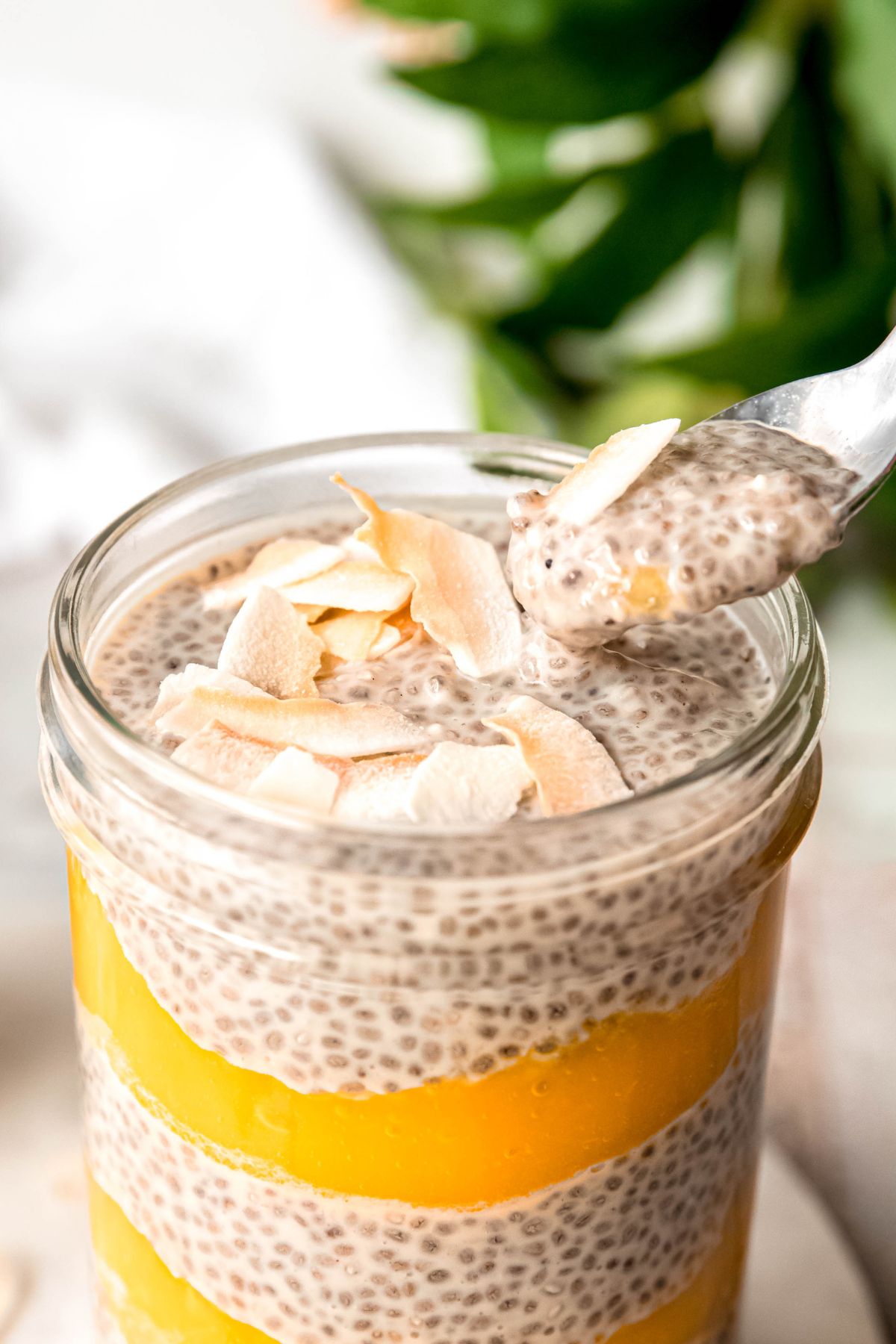silver spoon taking a bite of mango chia pudding parfait from the glass, showing the tapioca-like consistency of the chia seed layer and a hint of mango puree.