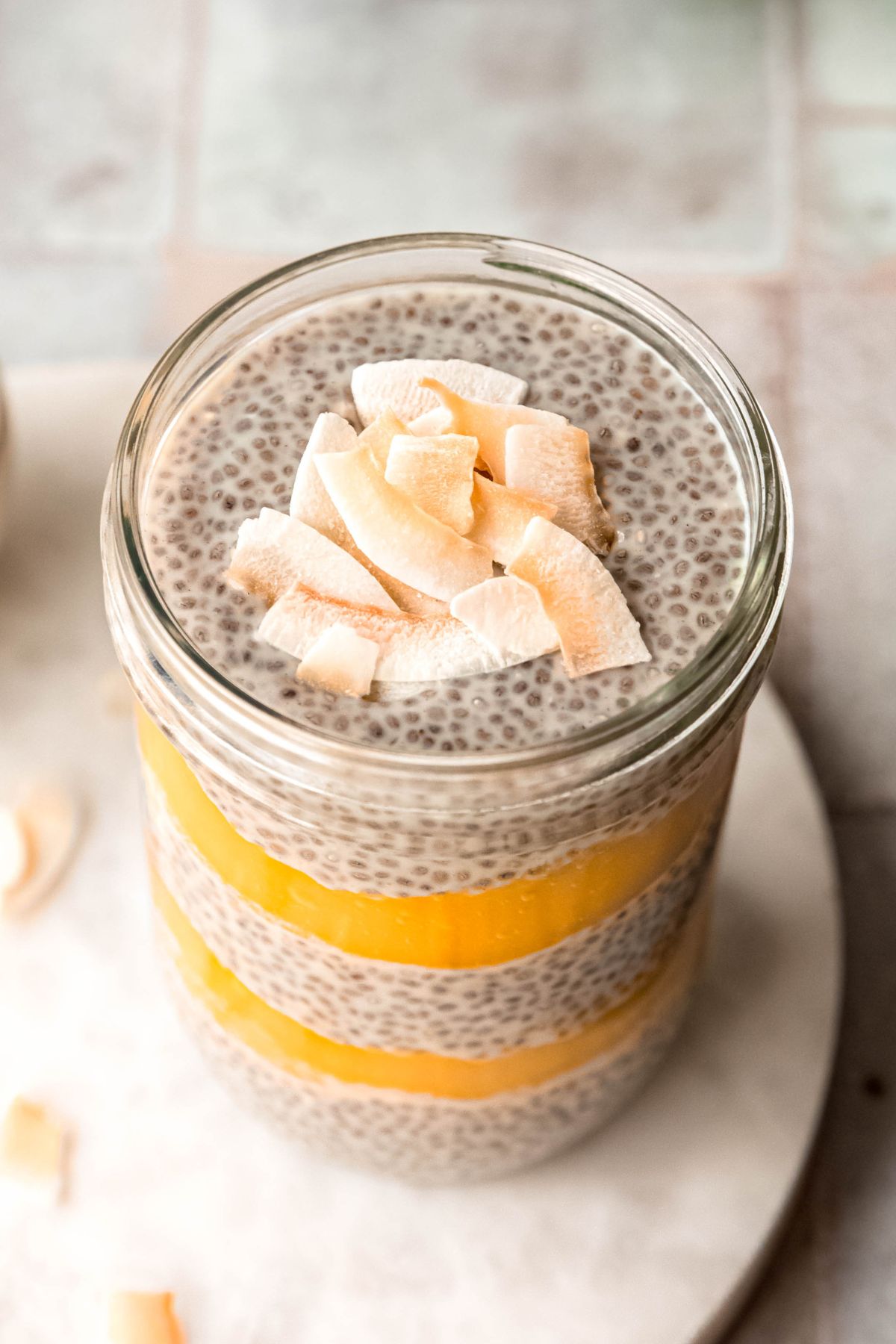 closeup 45 degree overhead shot looking down on a mason jar with mango chia pudding assembled as a parfait with layers of creamy coconut chia pudding and bright mango puree with a pudding layer ending the layers and a sprinkle of toasted coconut flakes for garnish.