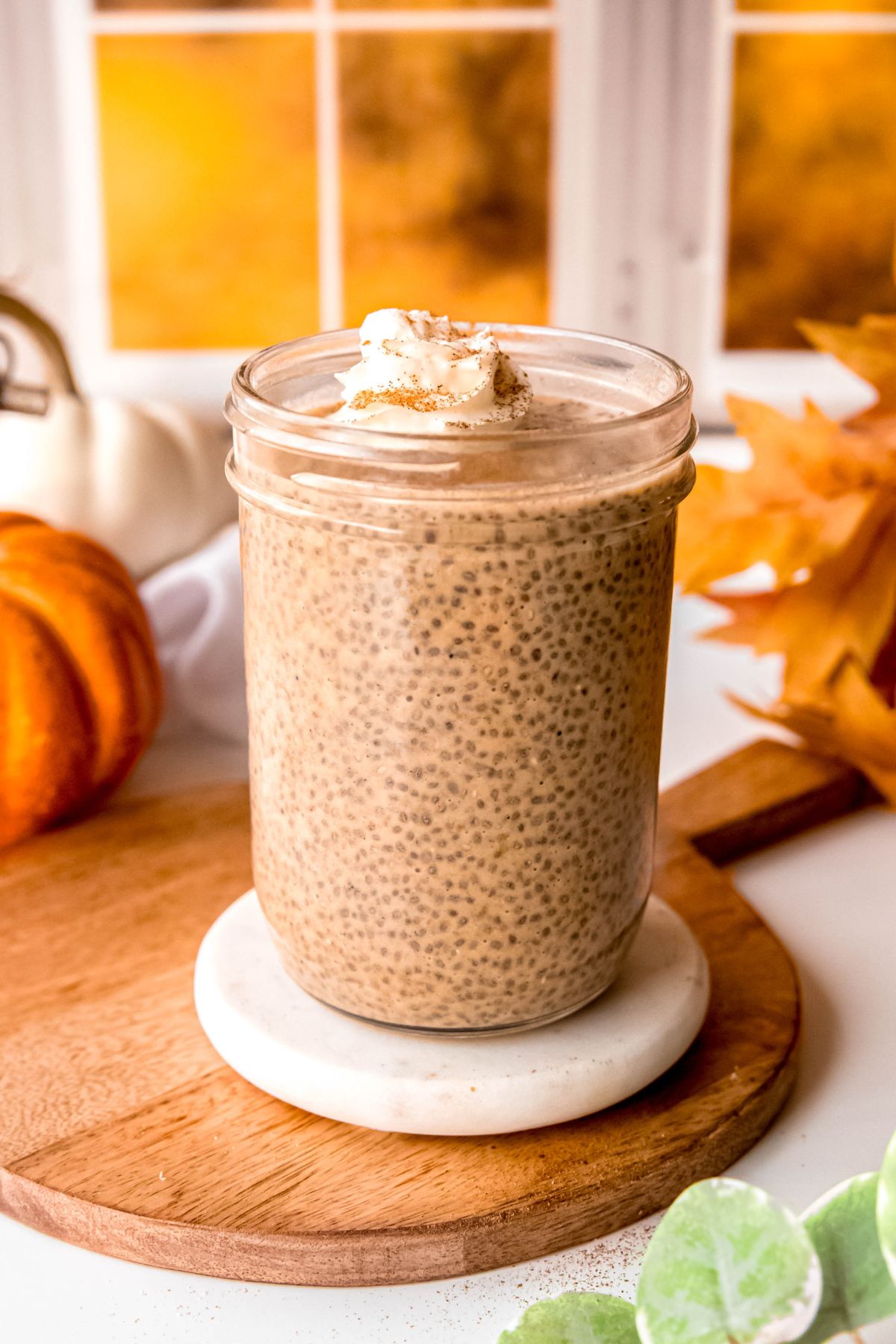 mason jar of pumpkin pie chia pudding on a table with orange and white pumpkins, a bouquet of fall leaves, and a green houseplant.