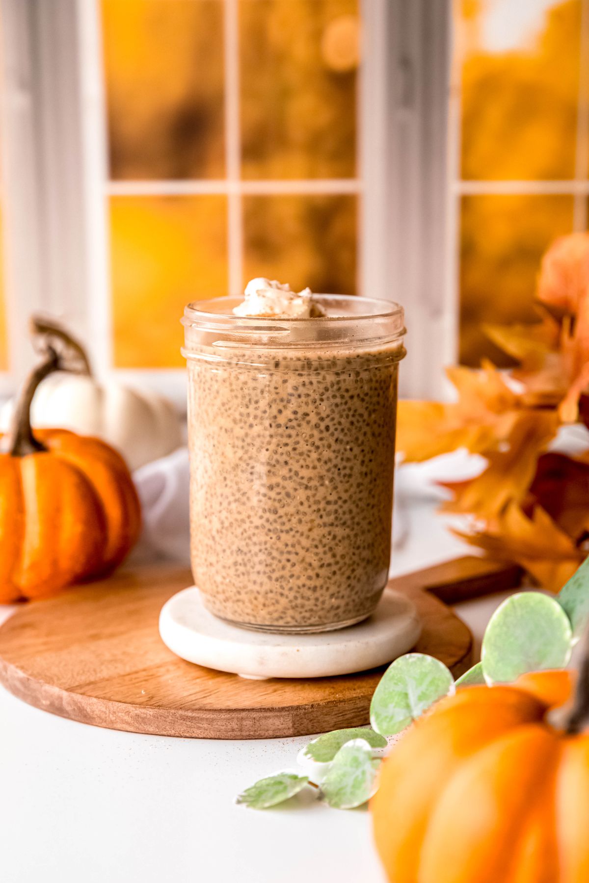 pint sized jar of pumpkin chia pudding on a wooden board atop a white table decorated with white and orange pumpkins, a bouquet of fall leaves and a green houseplant.