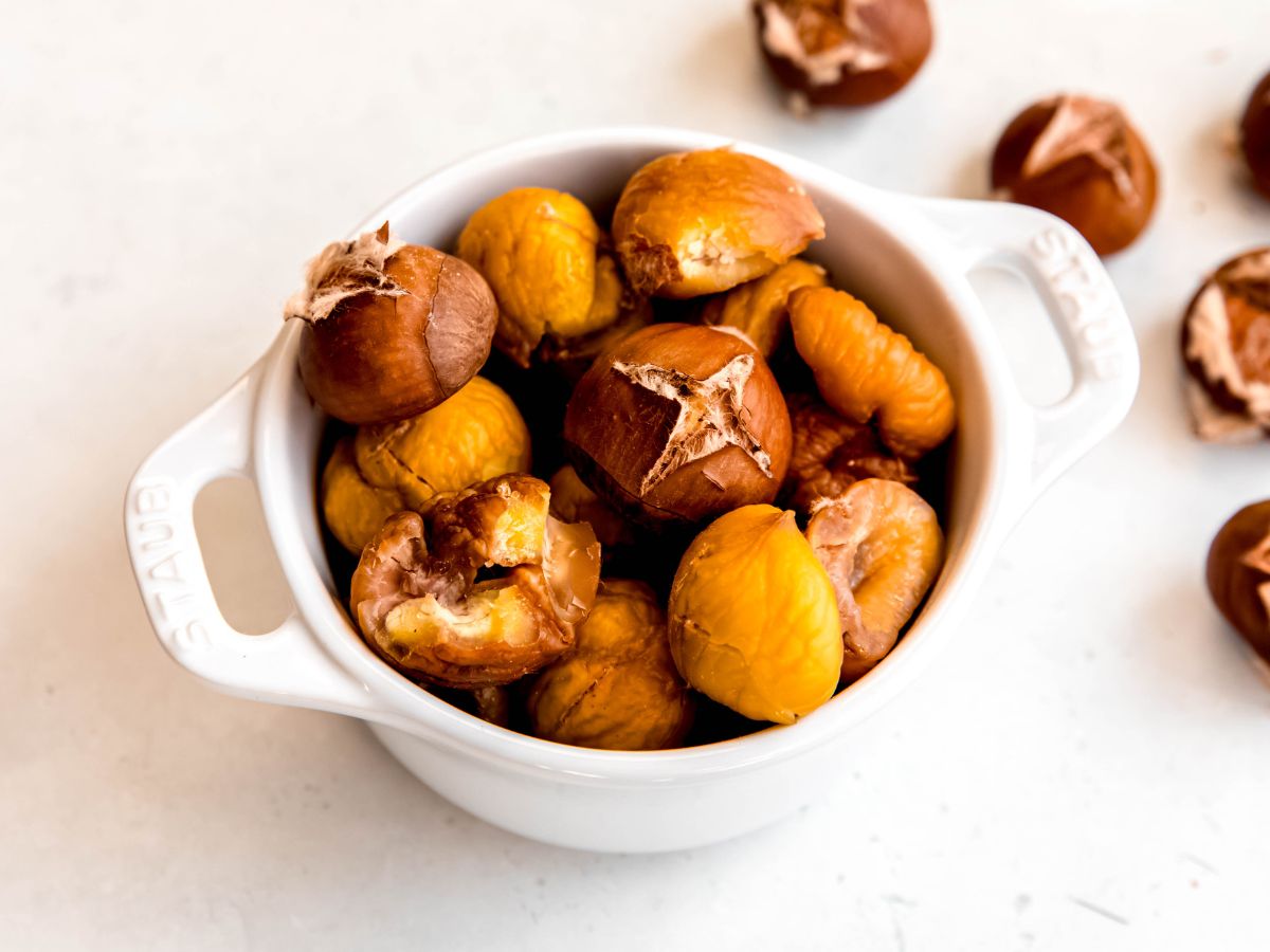 horizontally framed shot of a small white staub bowl filled with peeled roasted chestnuts on a white table with some unpeeled roasted chestnuts scattered around.