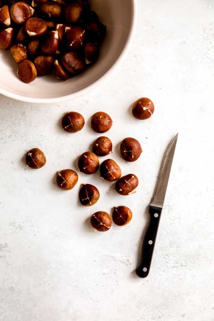 bowl of raw chestnuts with a handful that are on the work surface with a serrated knife after scoring.
