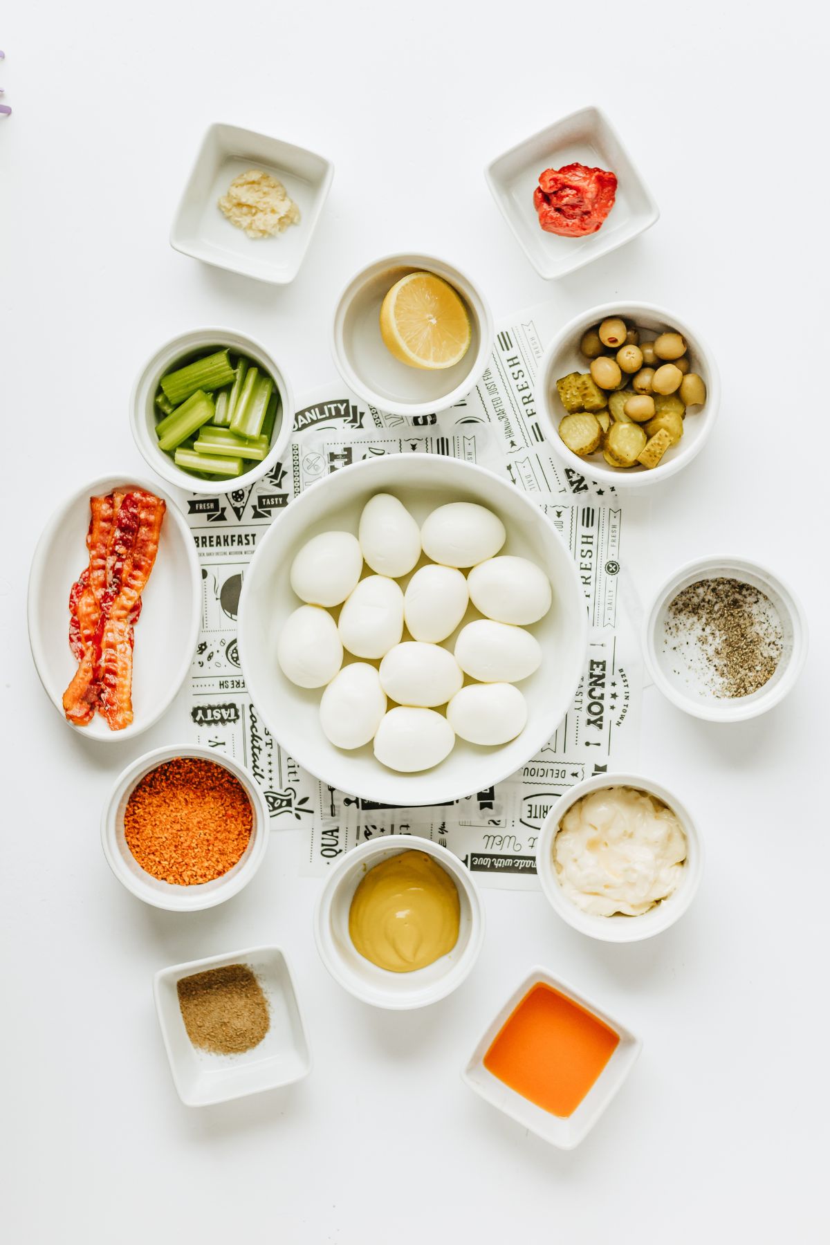 ingredients needed to make bloody mary deviled egg recipe measured out in white bowls on a white table with some food-inspired newspaper pieces beneath the bowl of eggs.