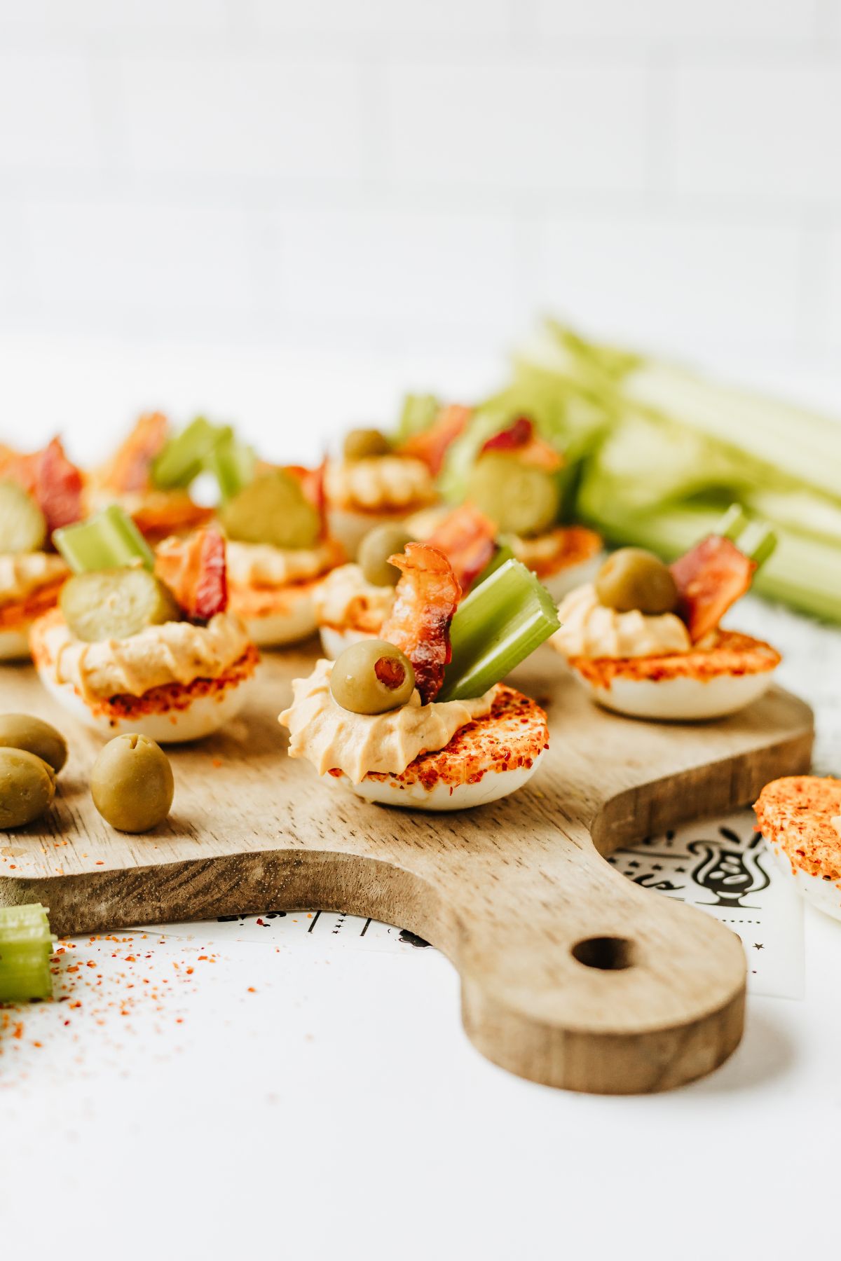 side on view of a wooden serving tray filled with bloody mary deviled eggs on a white table with more celery sticks and a sprinkle of tajin.