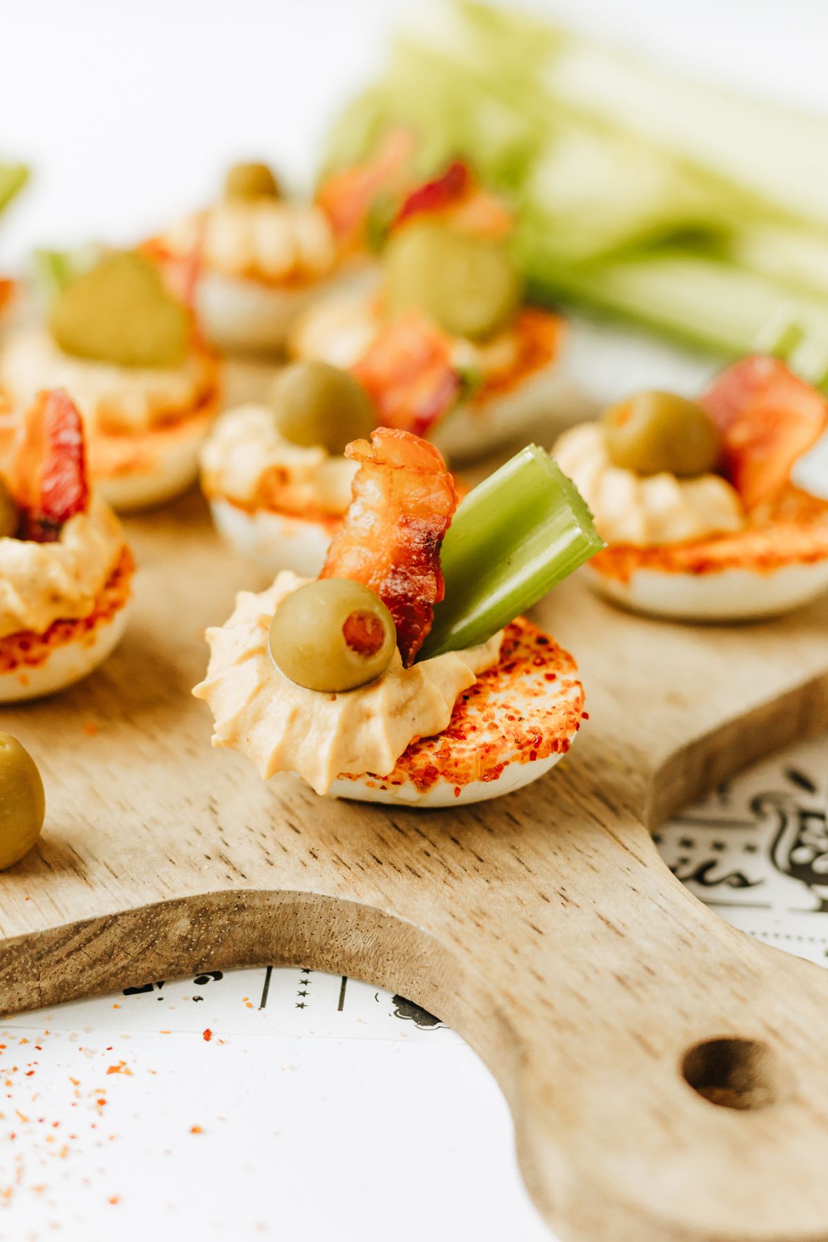 closeup shot of a wooden serving tray with the nearest bloody mary deviled egg in focus with the rest blurred behind; it's garnished with bacon, celery, and a cocktail olive.