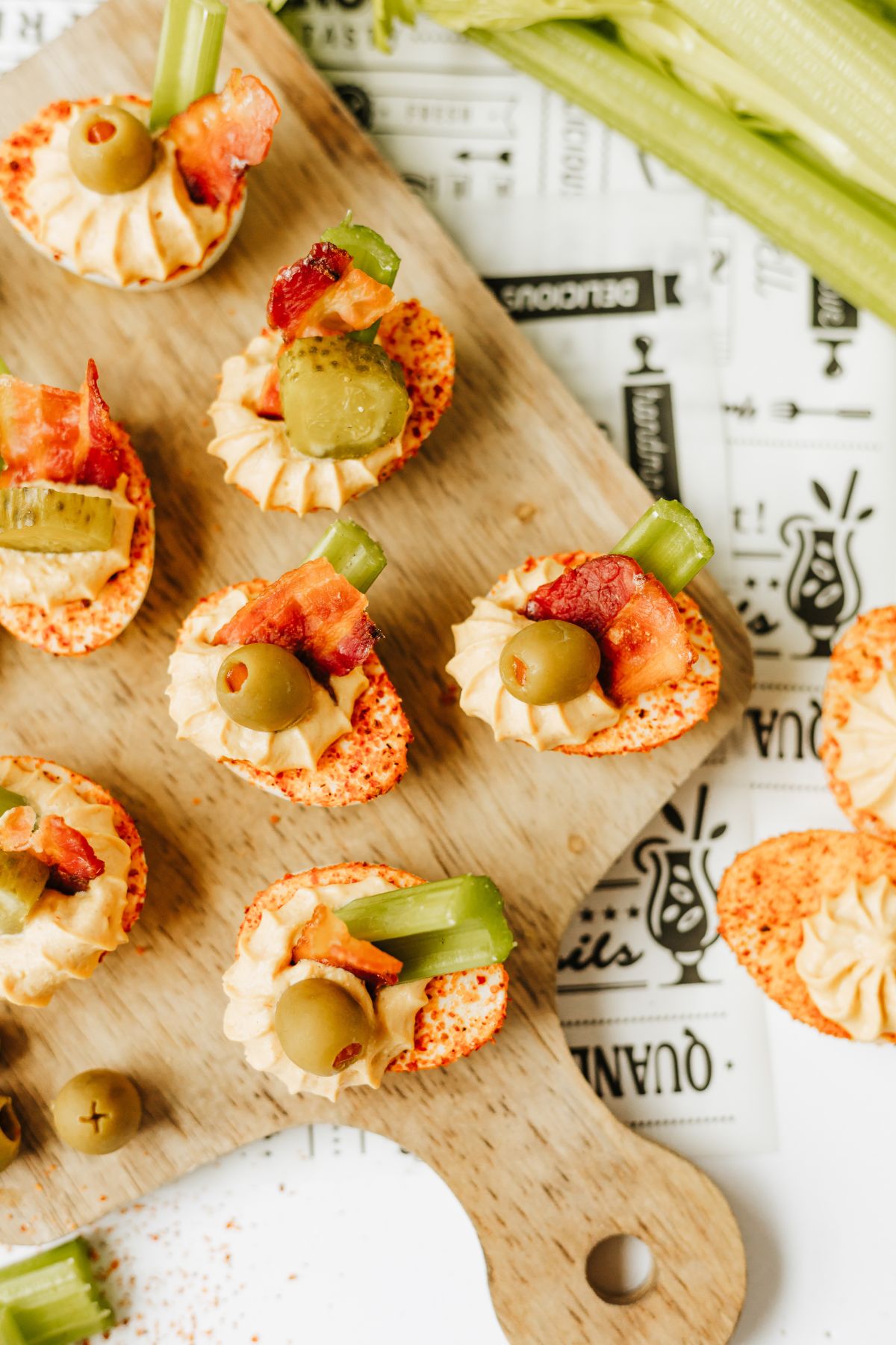 overhead shot of a wooden serving tray filled with bloody mary deviled eggs garnished with bacon, cocotail olives or pickle slices, and celery sticks.