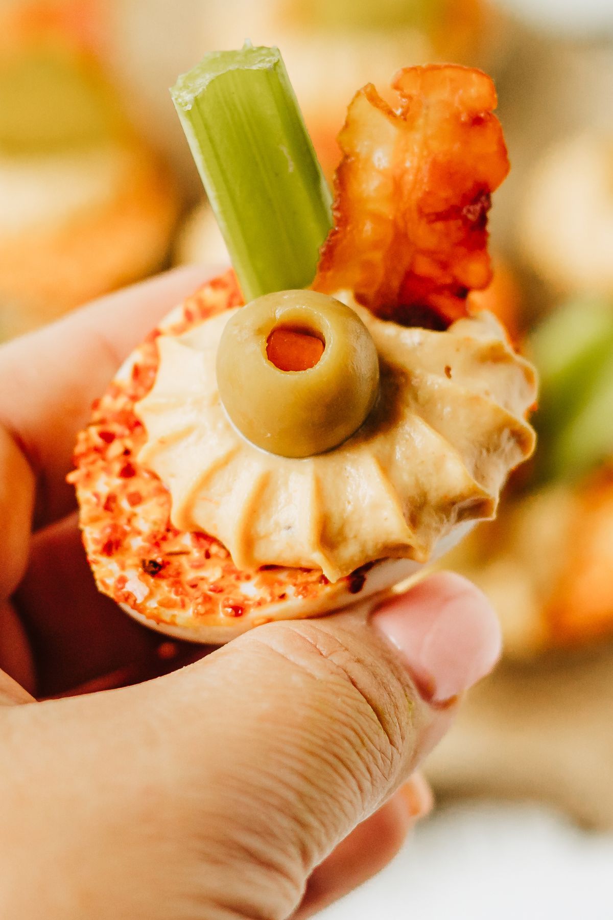 closeup shot of a hand holding a single bloody mary deviled egg showing the layers of red tajin, pale tan egg filling, a small cocktail olive, a short celery stick, and a small strip of crispy bacon.
