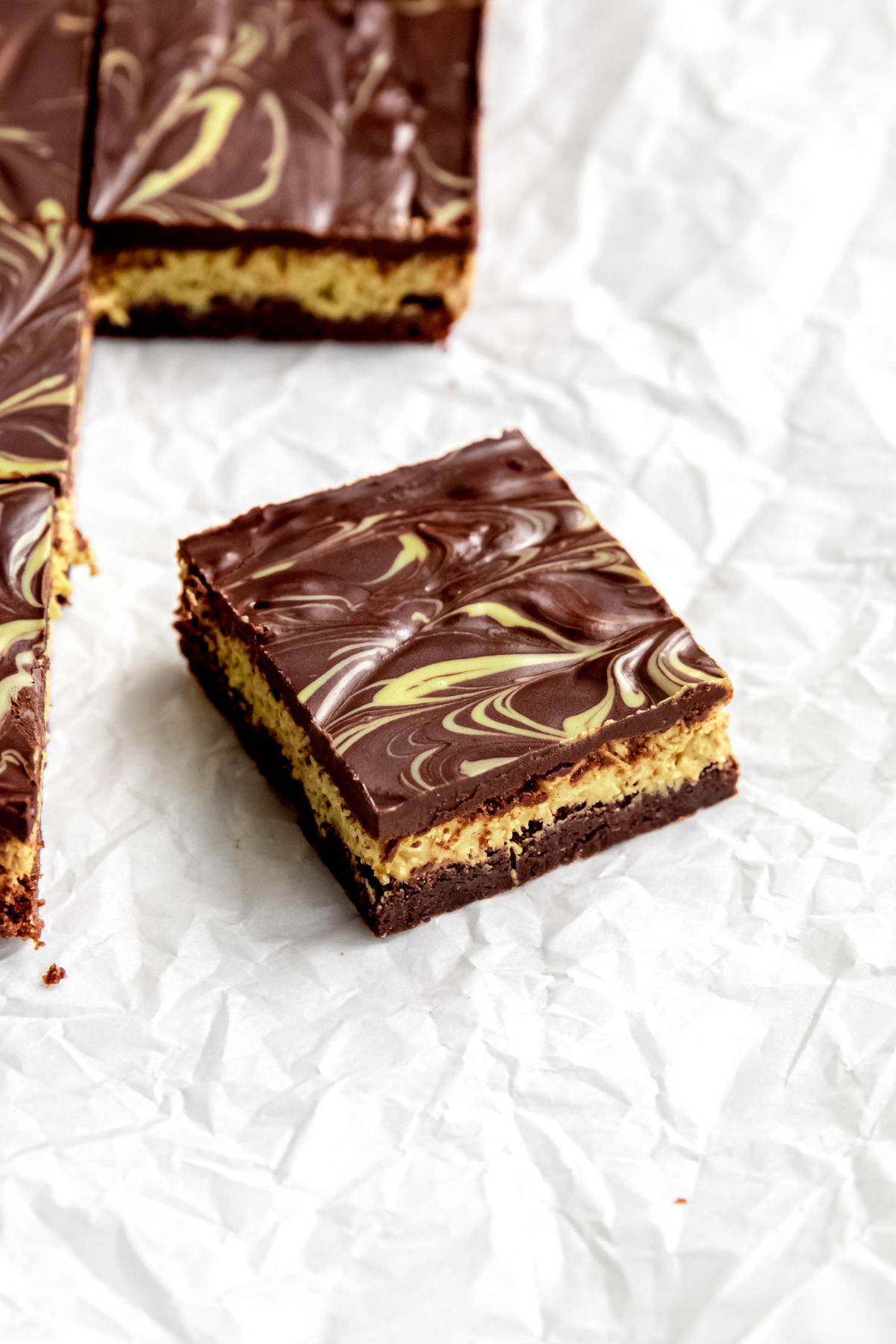 closeup 45 degree angle shot of a dubai chocolate brownie on a piece of crumpled parchment paper; the brownie is turned on an angle and is showing the three distinct layers of brownie, pistachio filling, and chocolate ganache.