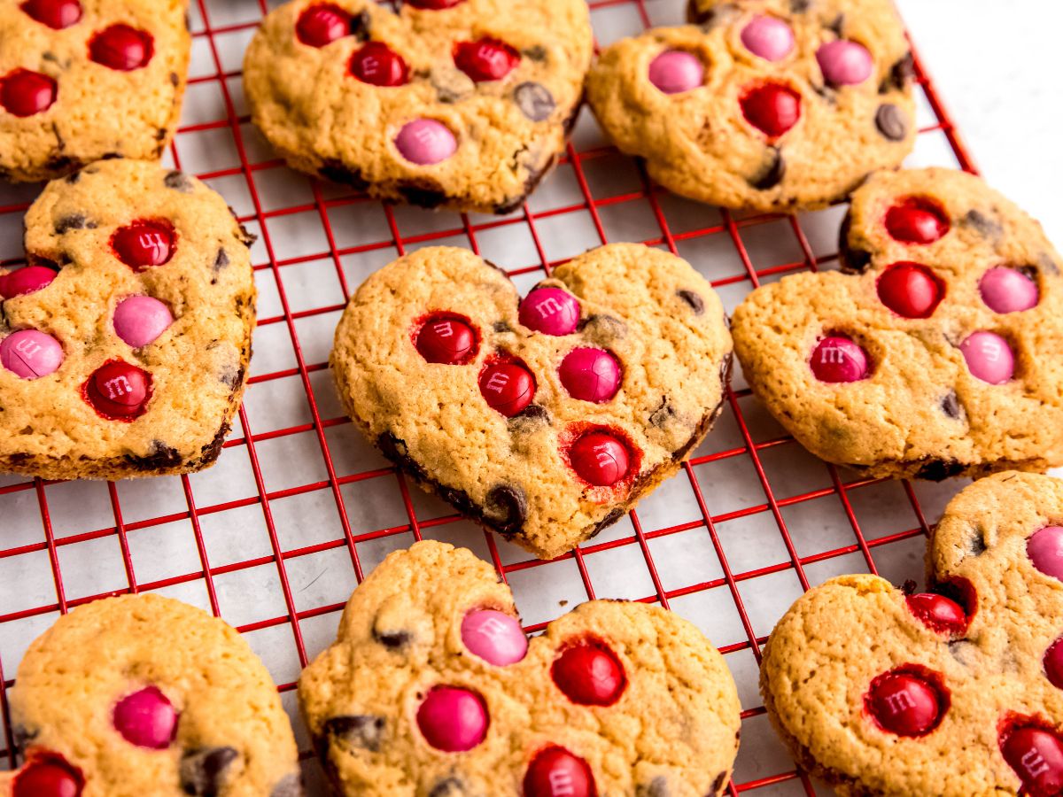 horizontally framed 45 degree angle closeup shot of festive heart-shaped chocolate chip cookies with valentine's day M&Ms on a red cooling rack.