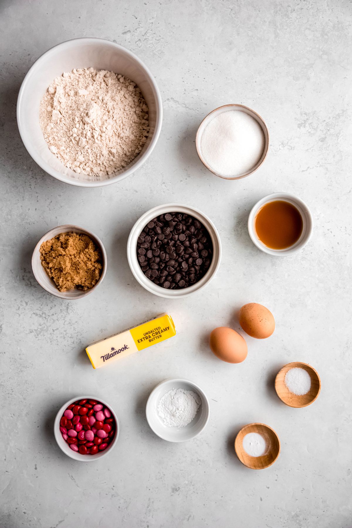 ingredients needed to make loaded heart shaped chocolate chip cookies with colorful M&Ms for valentine's day measured out on a white table.