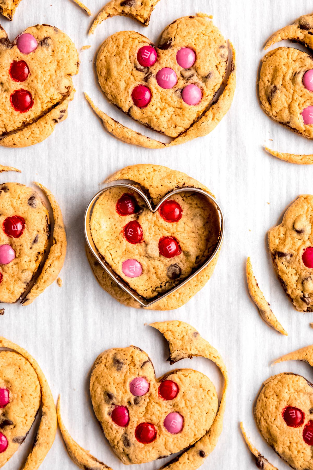 baked chocolate chip cookies being cut into heart shapes with a silver cookie cutter; there are some scraps from the cookies that are pulled away from the hearts.