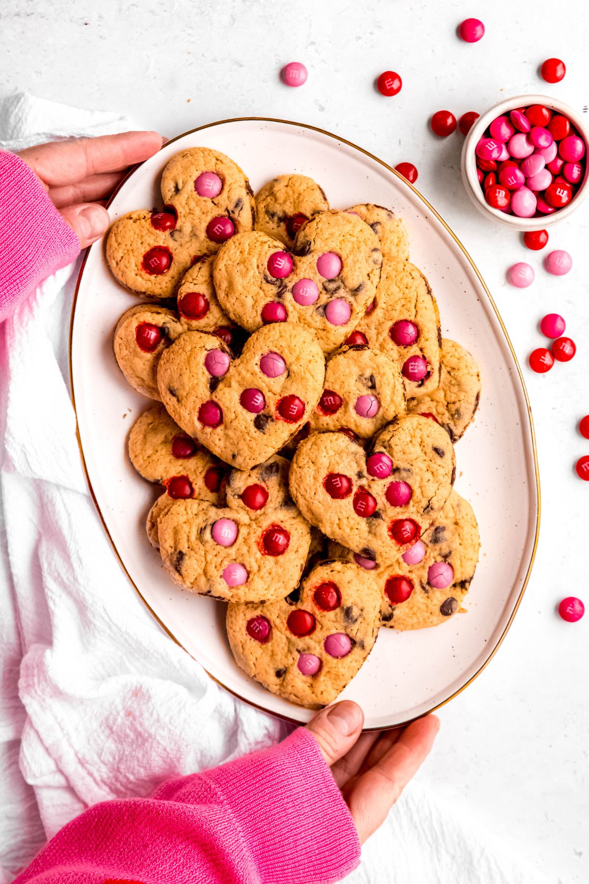 hands clad in a pink sweater up to the knuckles setting down an oval serving tray filled with heart shaped chocolate chip cookies for valnetines day on a white table with more M&Ms.