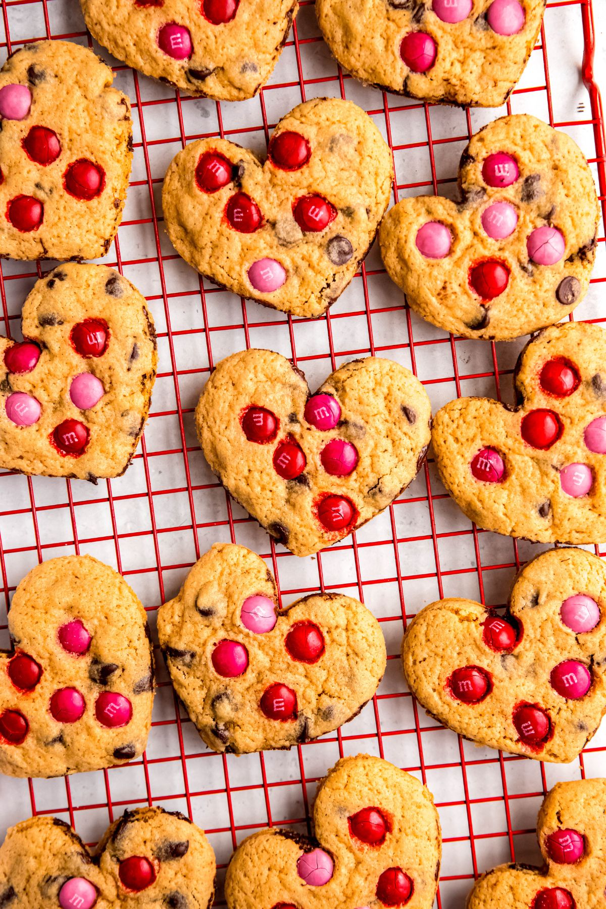 heart shaped chocolate chip cookies with red and pink M&Ms on a red cooling tray on a white table.