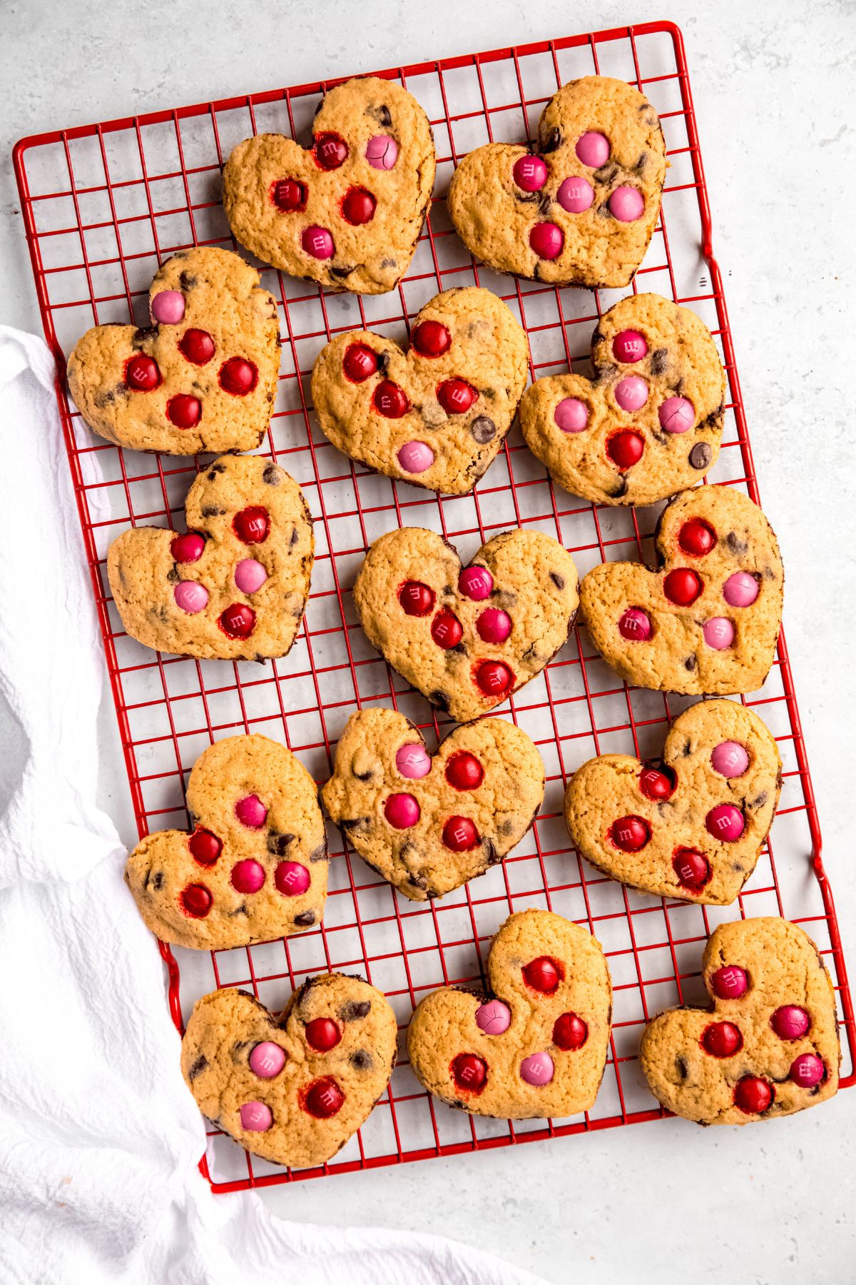 heart shaped chocolate chip cookies with red and pink M&Ms on a red cooling rack on a white table with a white linen cloth.