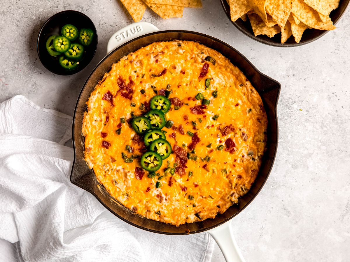 horizontally framed shot of a skillet of bacon jalapeno popper dip on a grey table with a black bowl of tortilla chips and a black pinch bowl of extra jalapeño slices.