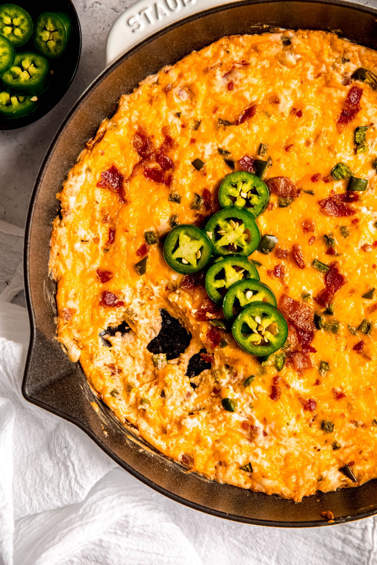 closeup overhead shot of a skillet of jalapeño popper dip with bacon and cheddar cheese with part of the skillet scooped out and empty with a few tortilla chips scattered around.