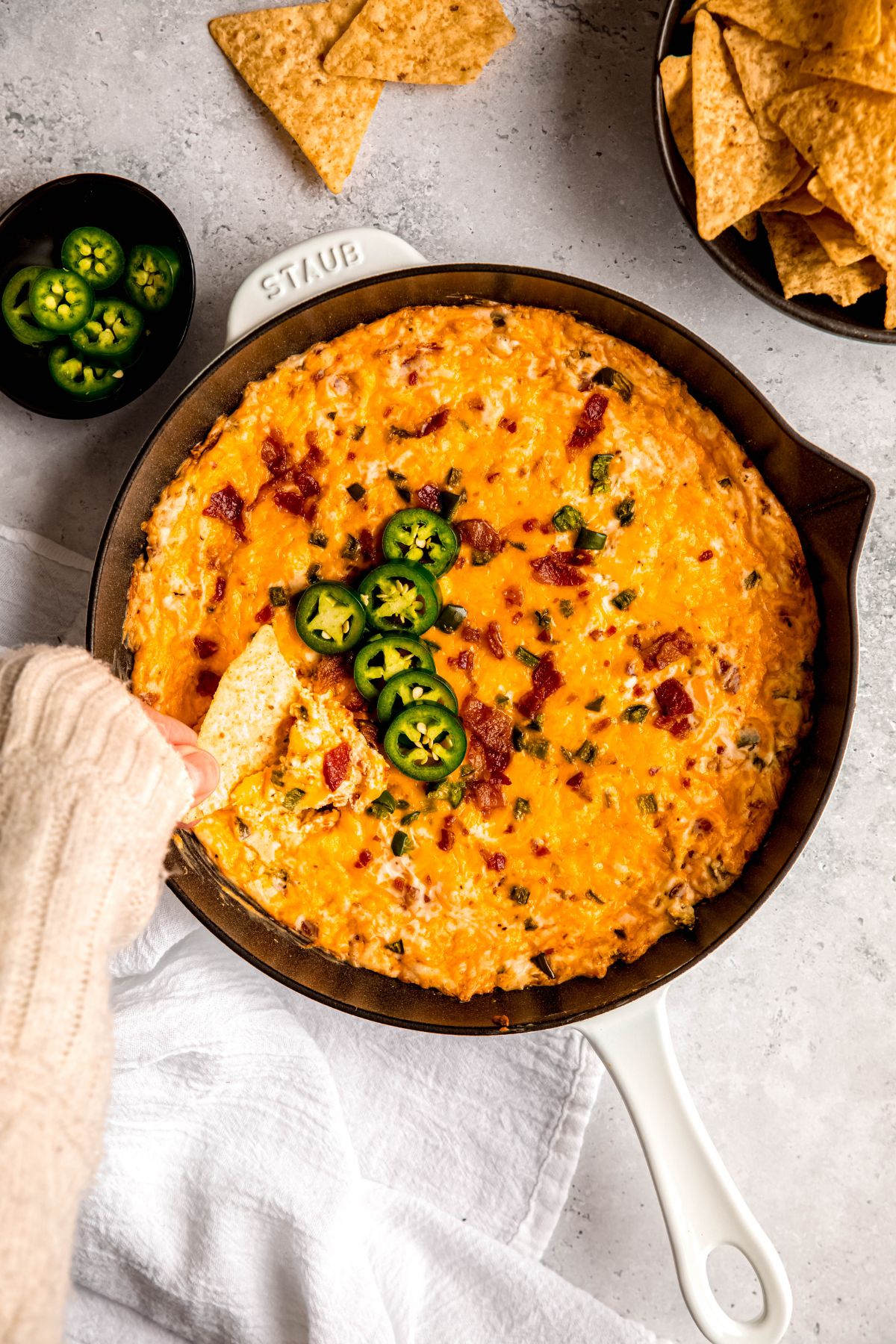 hand clad in a white sweater up to the knuckles dipping a tortilla chip into a melty skillet of creamy jalapeño popper dip.