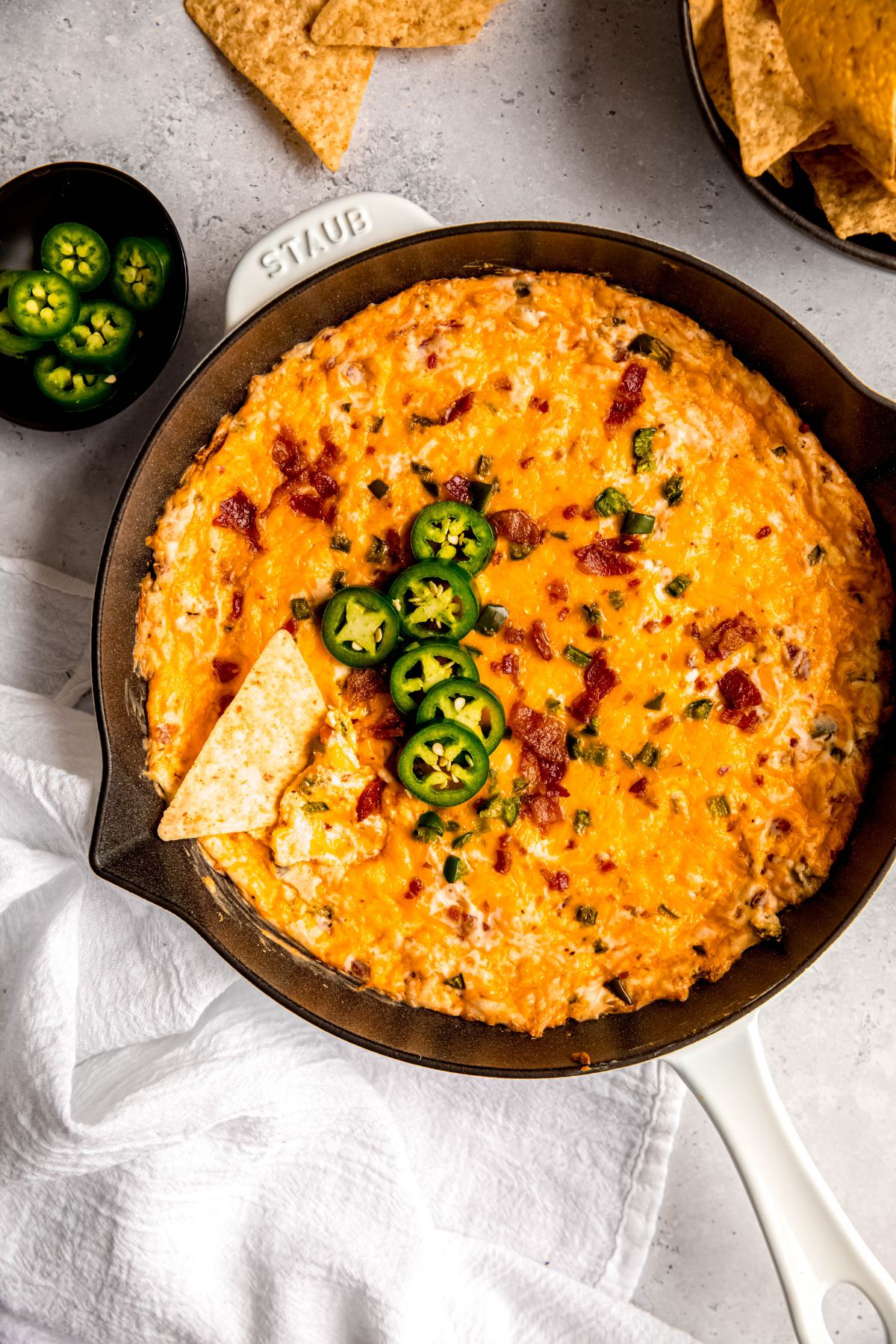 overhead shot of a white enameled cast iron skillet filled with easy jalapeño popper dip topped with fresh slices of jalapenos and a tortilla chip dug in.