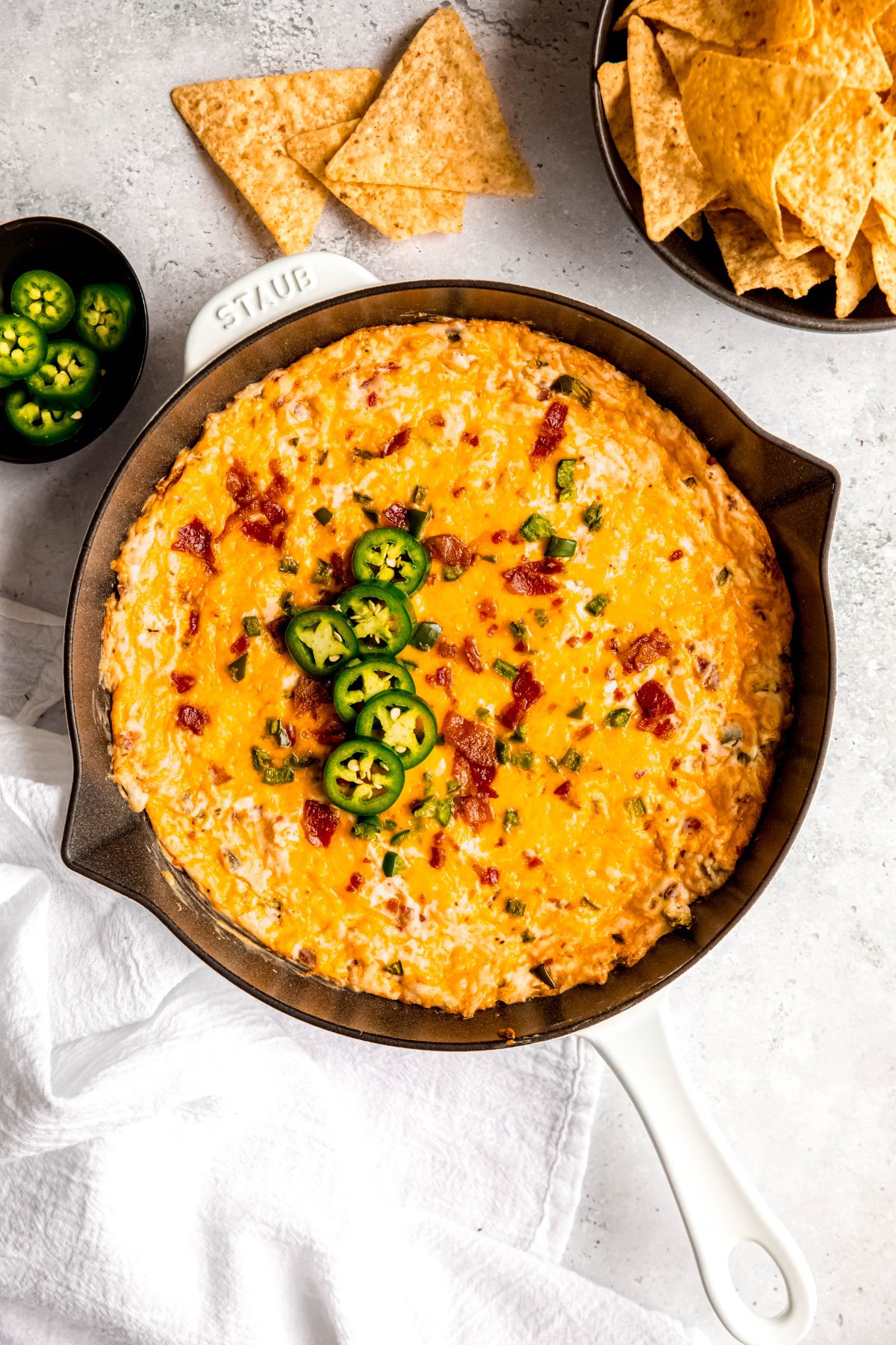 overhead shot of a skillet filled with baked jalapeño popper dip topped with fresh jalapeno slices on a white table with a bowl of tortilla chips and a pinch bowl with extra jalapeños.