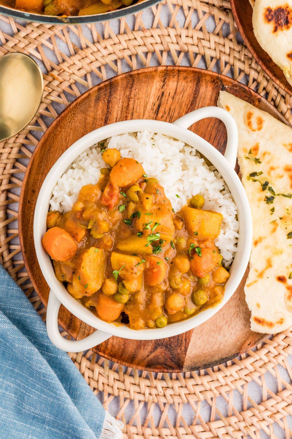 flat lay shot of a white soup bowl with handles filled with white rice and chickpea potato curry, garnished with cilantro, on a wooden charger with a side of naan on top of a woven wooden placemat.