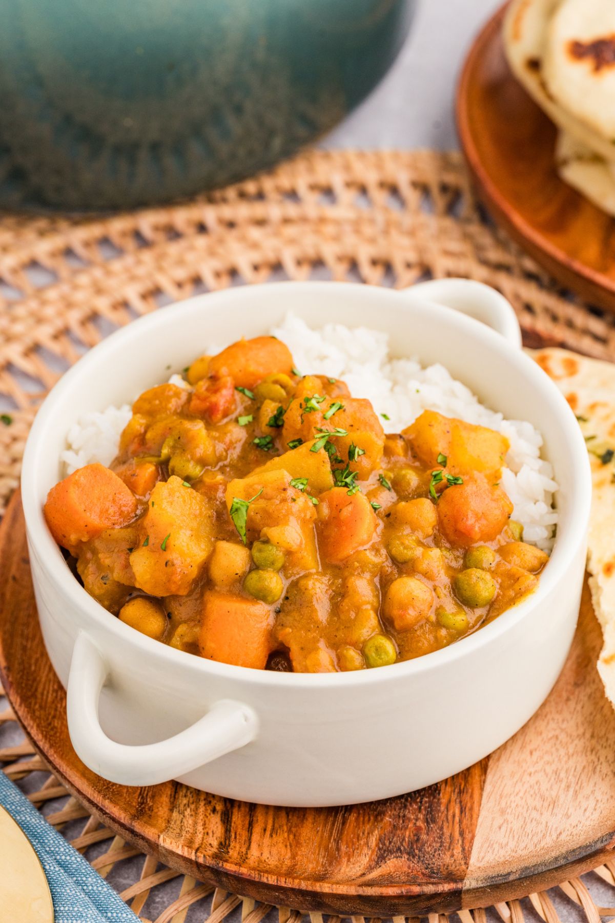 chickpea pistachio curry served over a bed of steamed rice in a white bowl with handles on a wooden charger plate with a piece of naan.