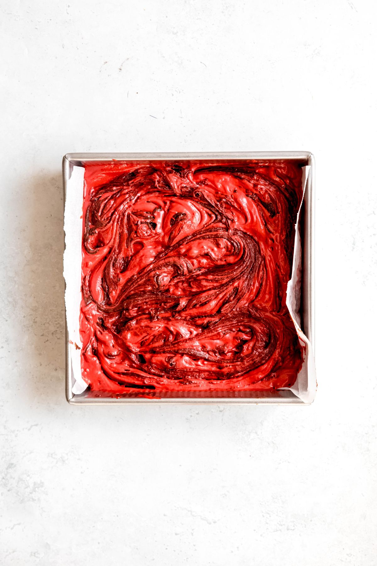 raw red velvet cheesecake brownies in the tin after swirling the brownie and cheesecake layers together.