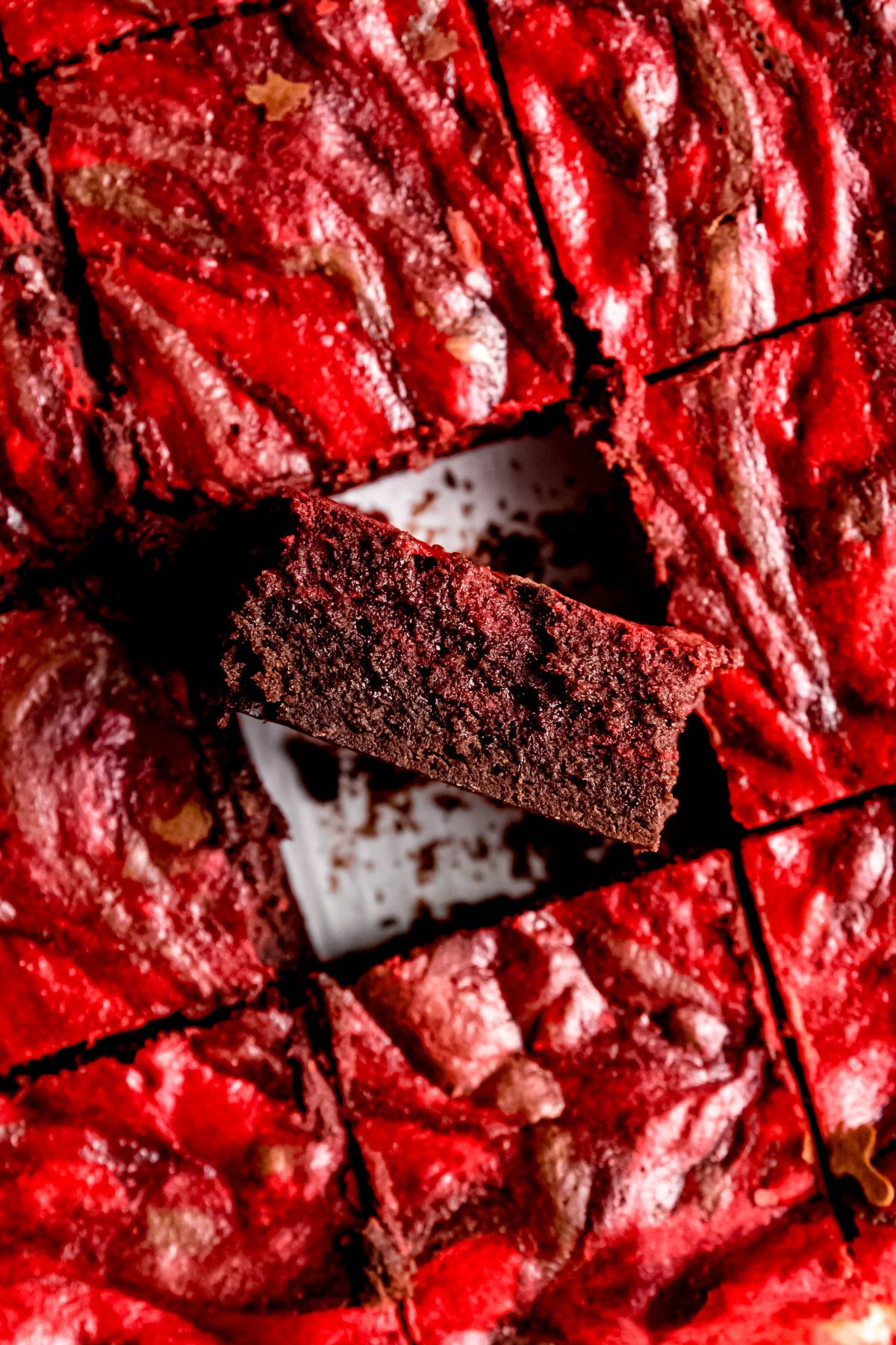 flat lay closeup shot of a tray of red velvet brownies with the one in the center turned on its side to show the fudgy consistency contrasting with the red cheesecake swirl on top.