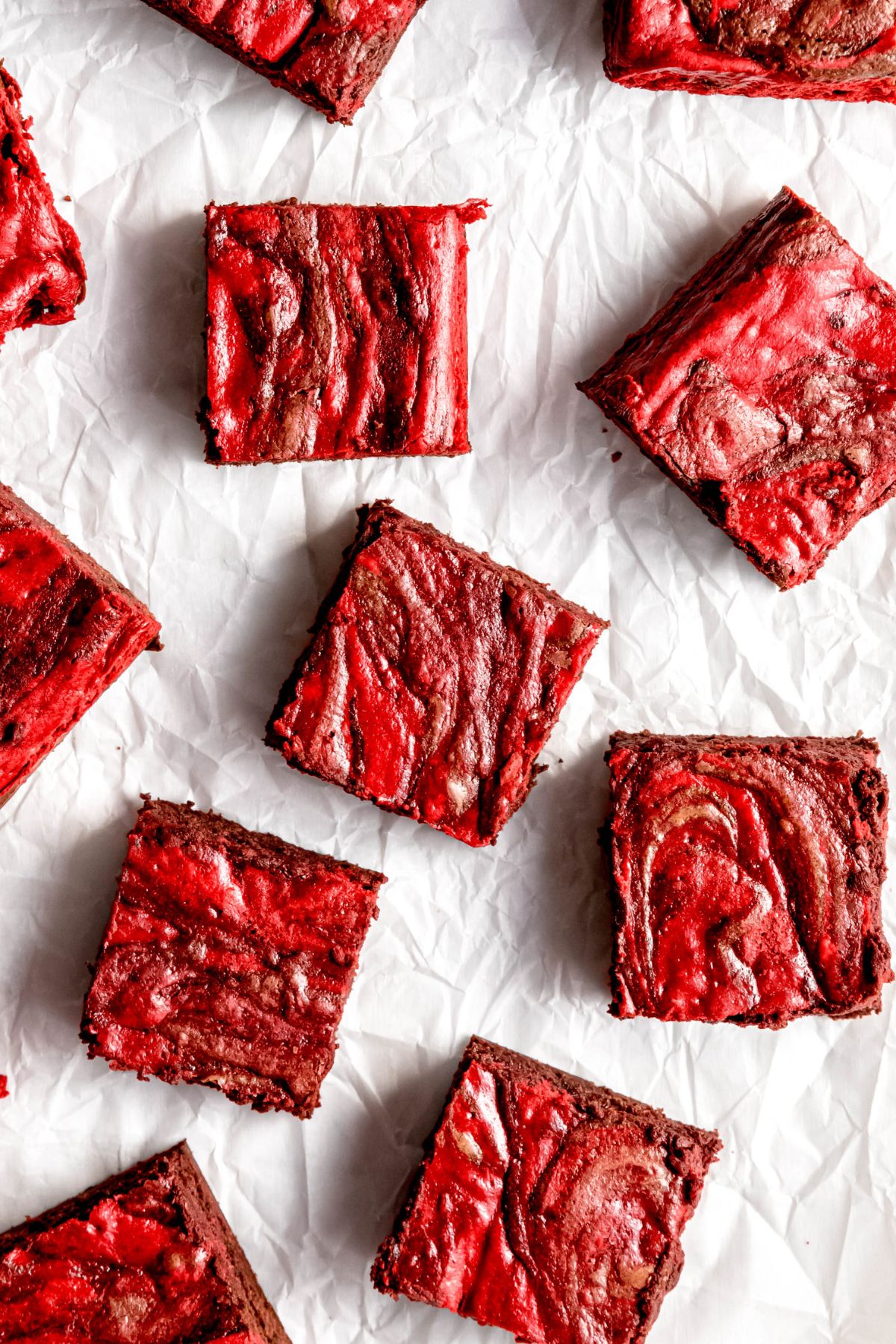 overhead shot of red velvet cheesecake brownies that have been cut into squares and scattered topsy turvy on a piece of crumpled white parchment paper.